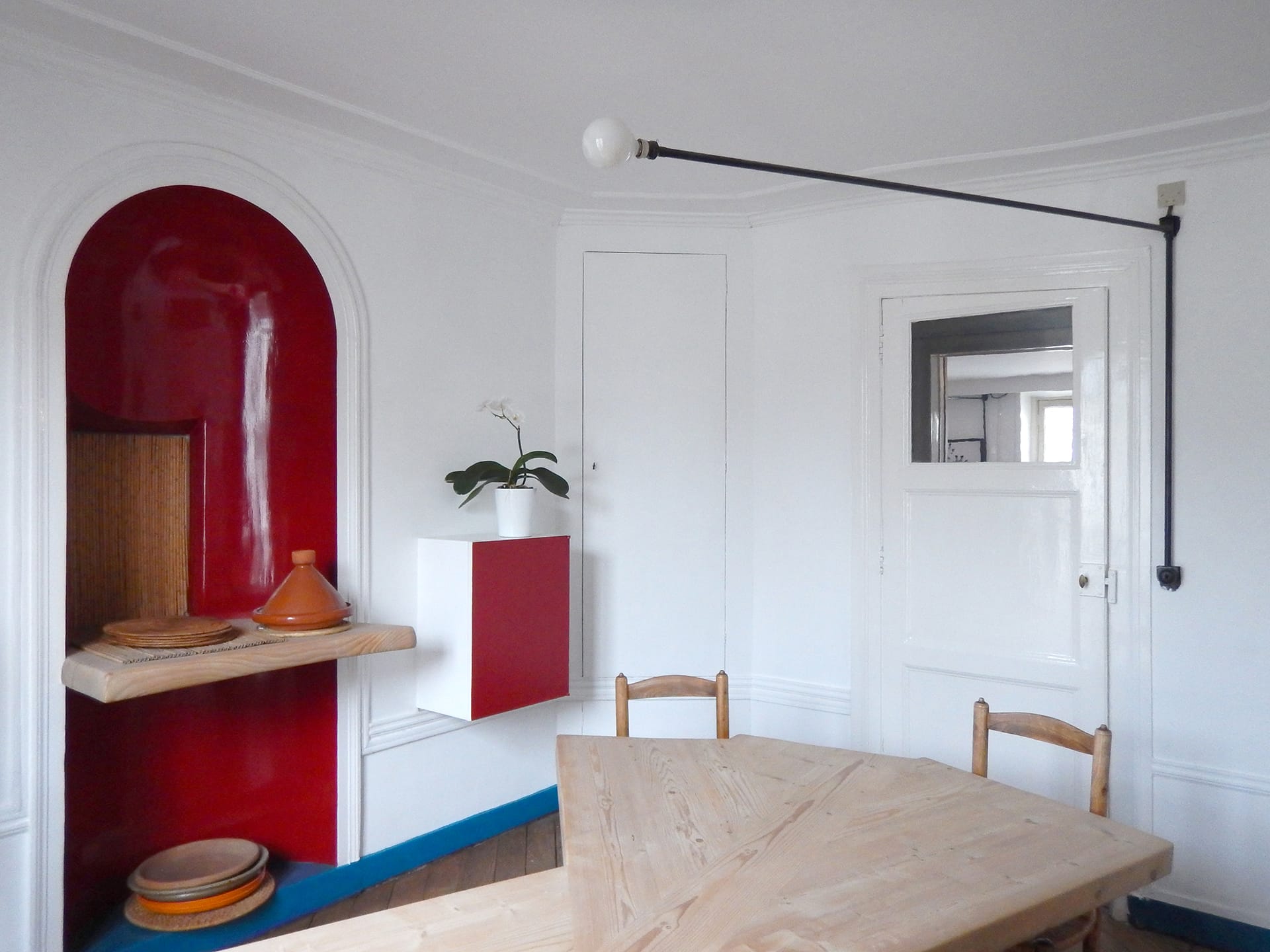 Minimalist dining room with white walls, red accents, a wooden table, chairs, and a wall-mounted light fixture.