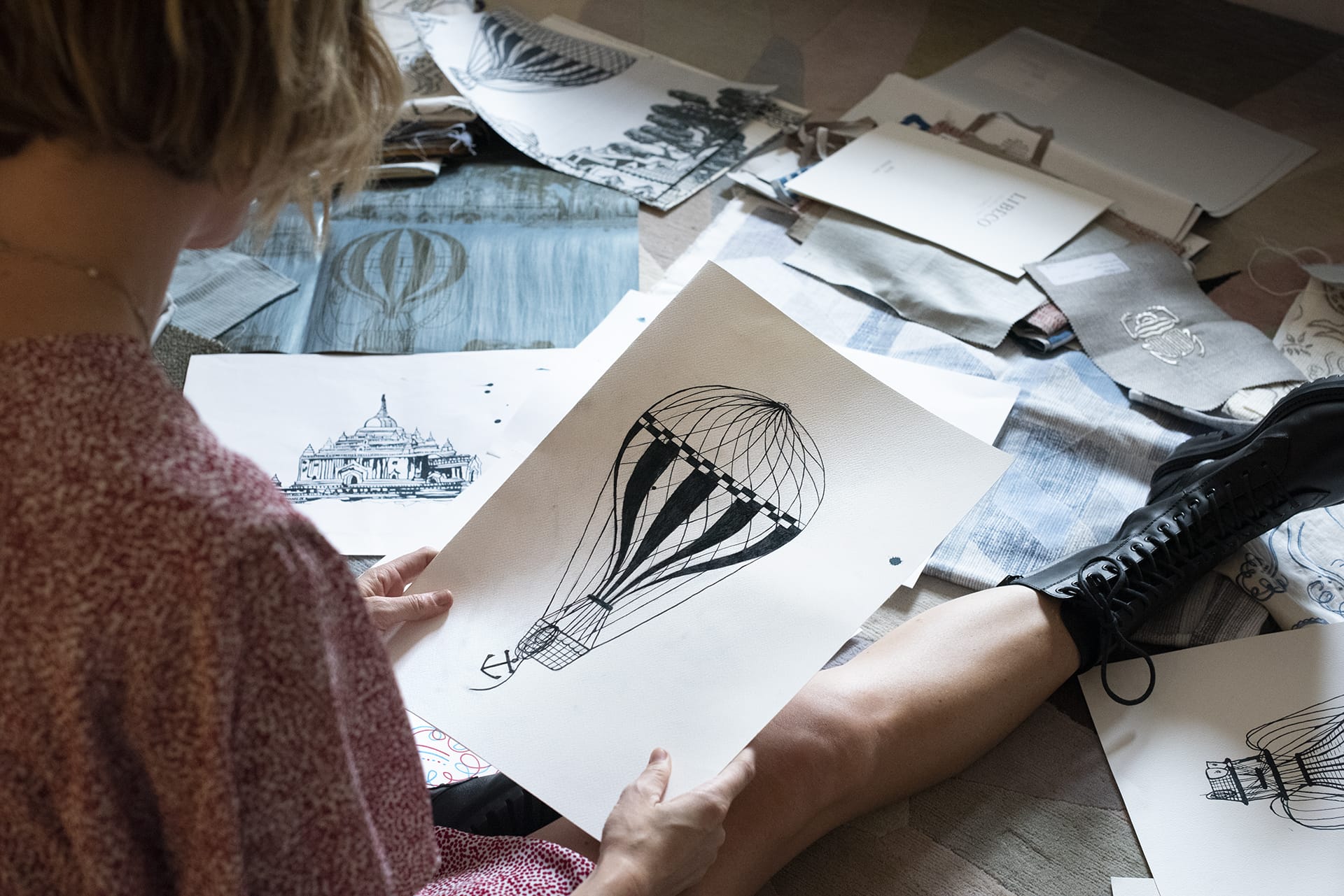 Person examining detailed pen drawing of a hot air balloon amidst an array of sketches and art supplies on the floor.