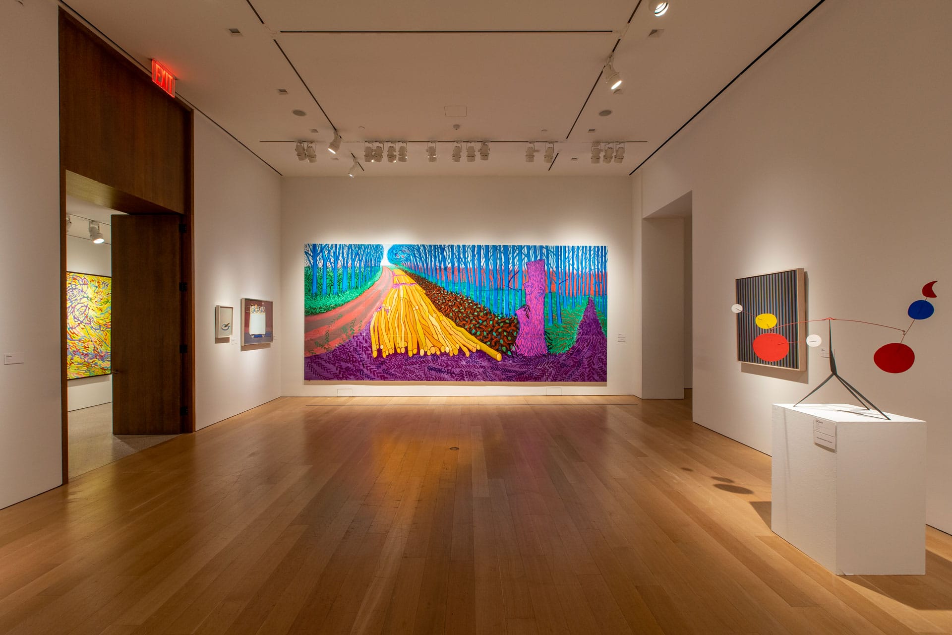 Art gallery with colorful abstract paintings and a metal sculpture on a pedestal in a well-lit room with wooden flooring.