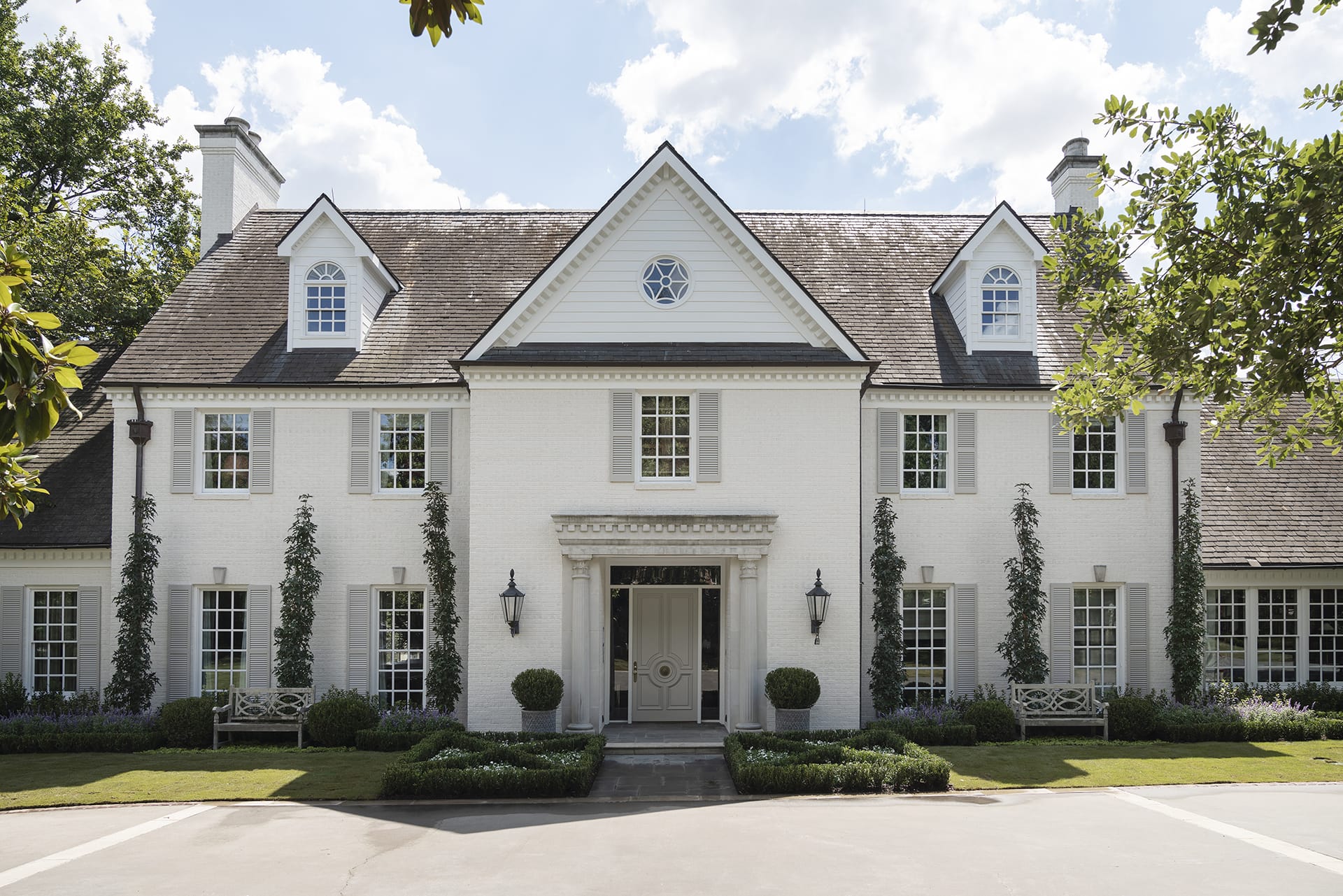Elegant two-story white brick house with symmetric windows, green shrubs, and a circular driveway under a blue sky.