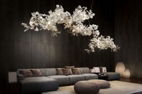 Modern living room with dark wood walls, gray sectional sofa, white floral chandeliers, wicker poufs, and soft lighting.