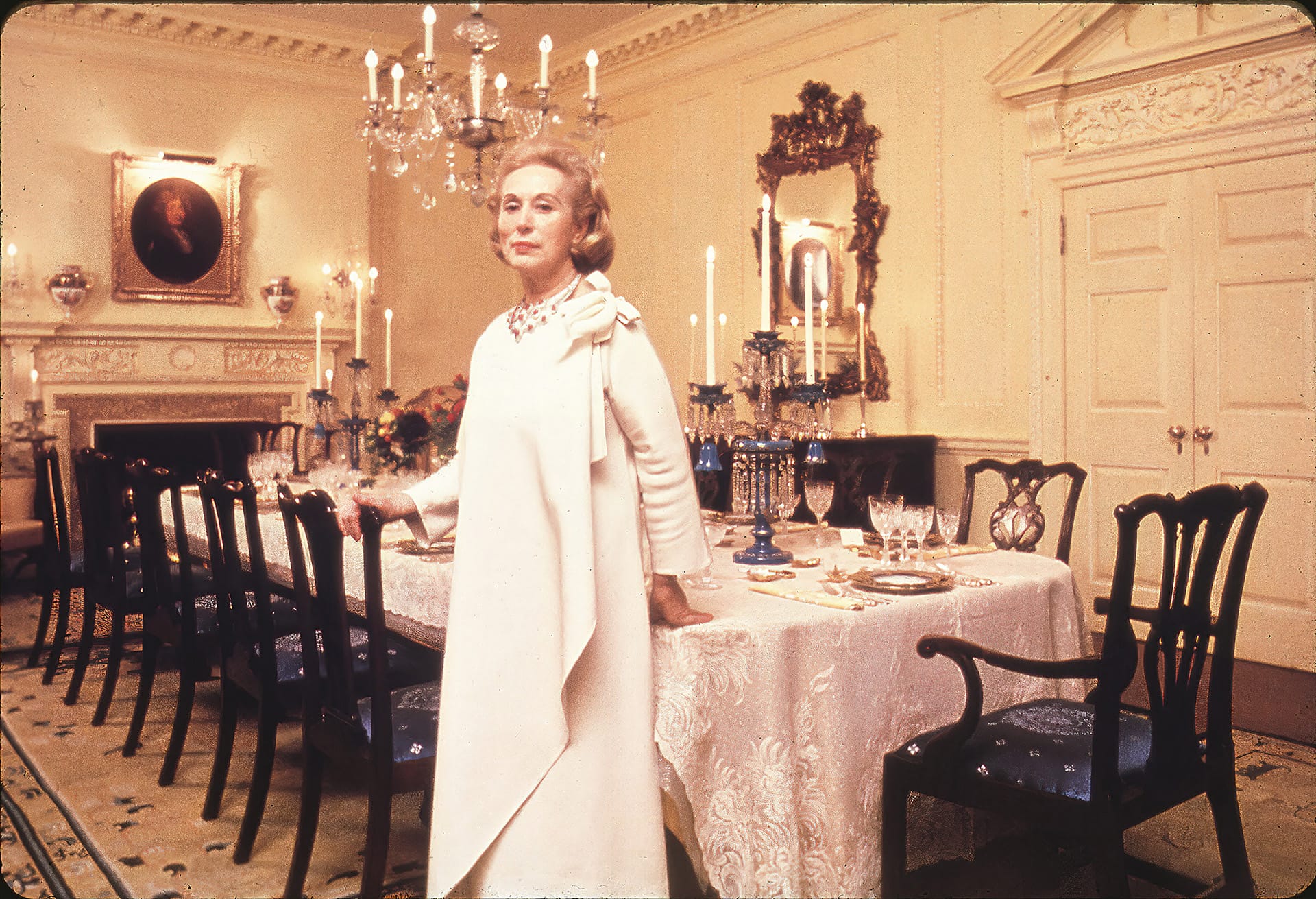 Elegant woman in white dress standing in luxurious dining room with ornate decor and set dining table.