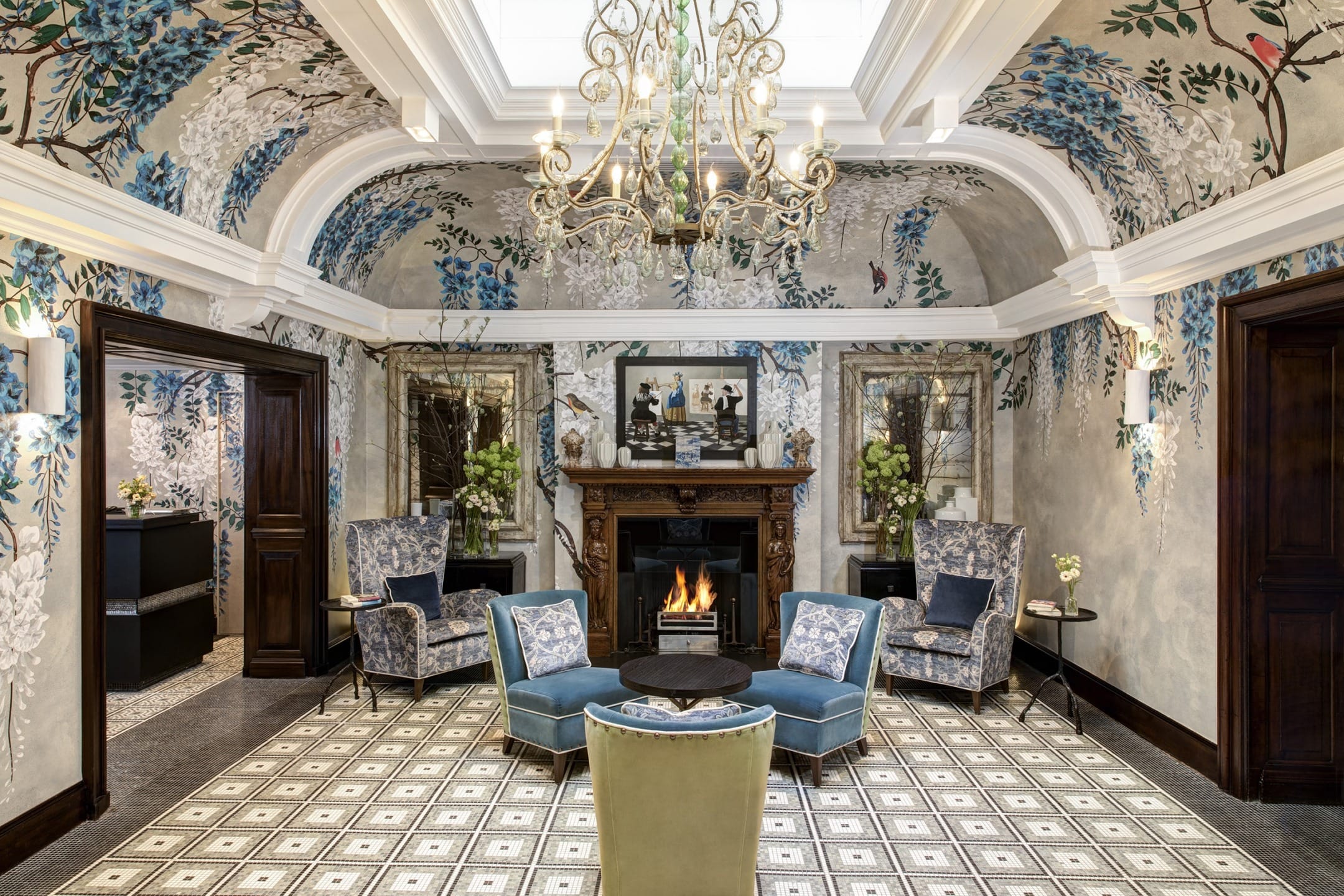 Elegant lounge with floral wallpaper, chandelier, fireplace, and blue armchairs around a central table on patterned floor.