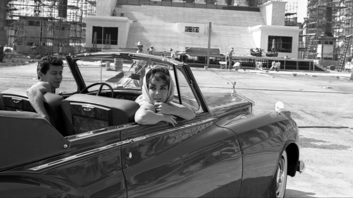 Vintage scene with a woman in a convertible, a man beside her, and a partially constructed building in the background.