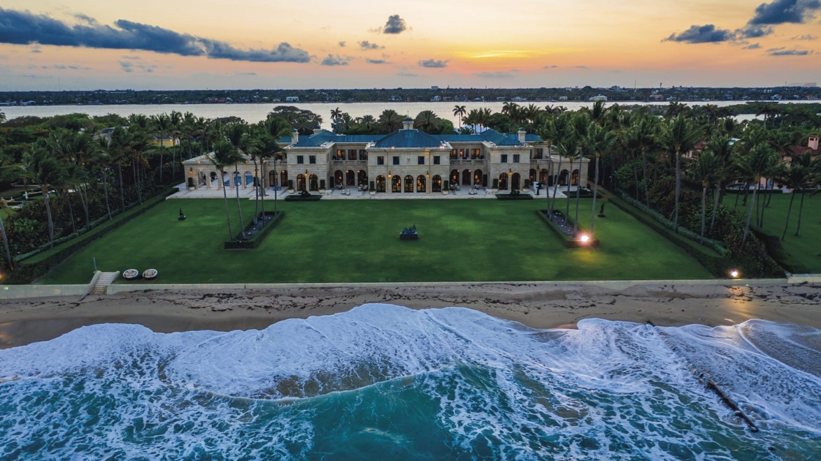 Luxurious beachfront mansion at sunset with palm trees and waves, overlooking a serene ocean view.
