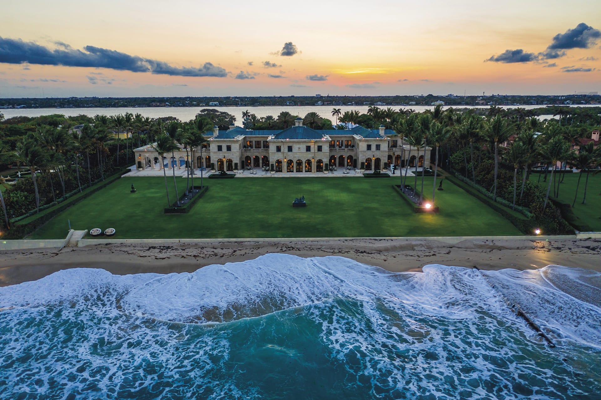 Luxurious beachfront mansion at sunset with palm trees and waves, overlooking a serene ocean view.