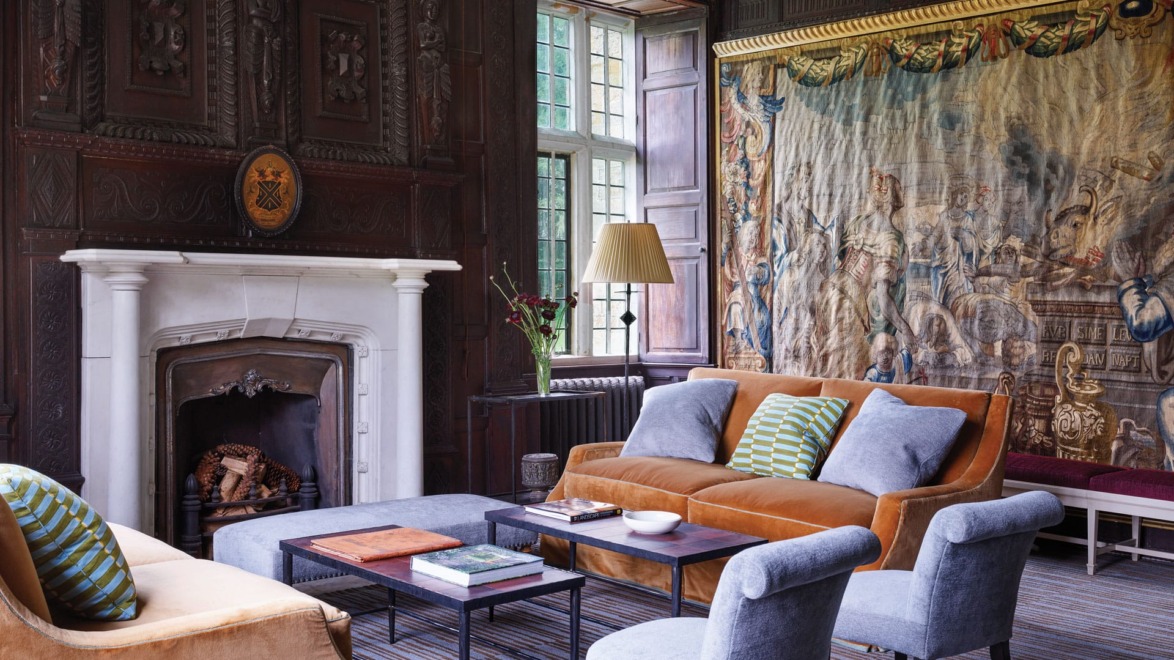 Elegant living room with vintage tapestry, fireplace, orange sofa, blue chairs, striped rug, and large window.