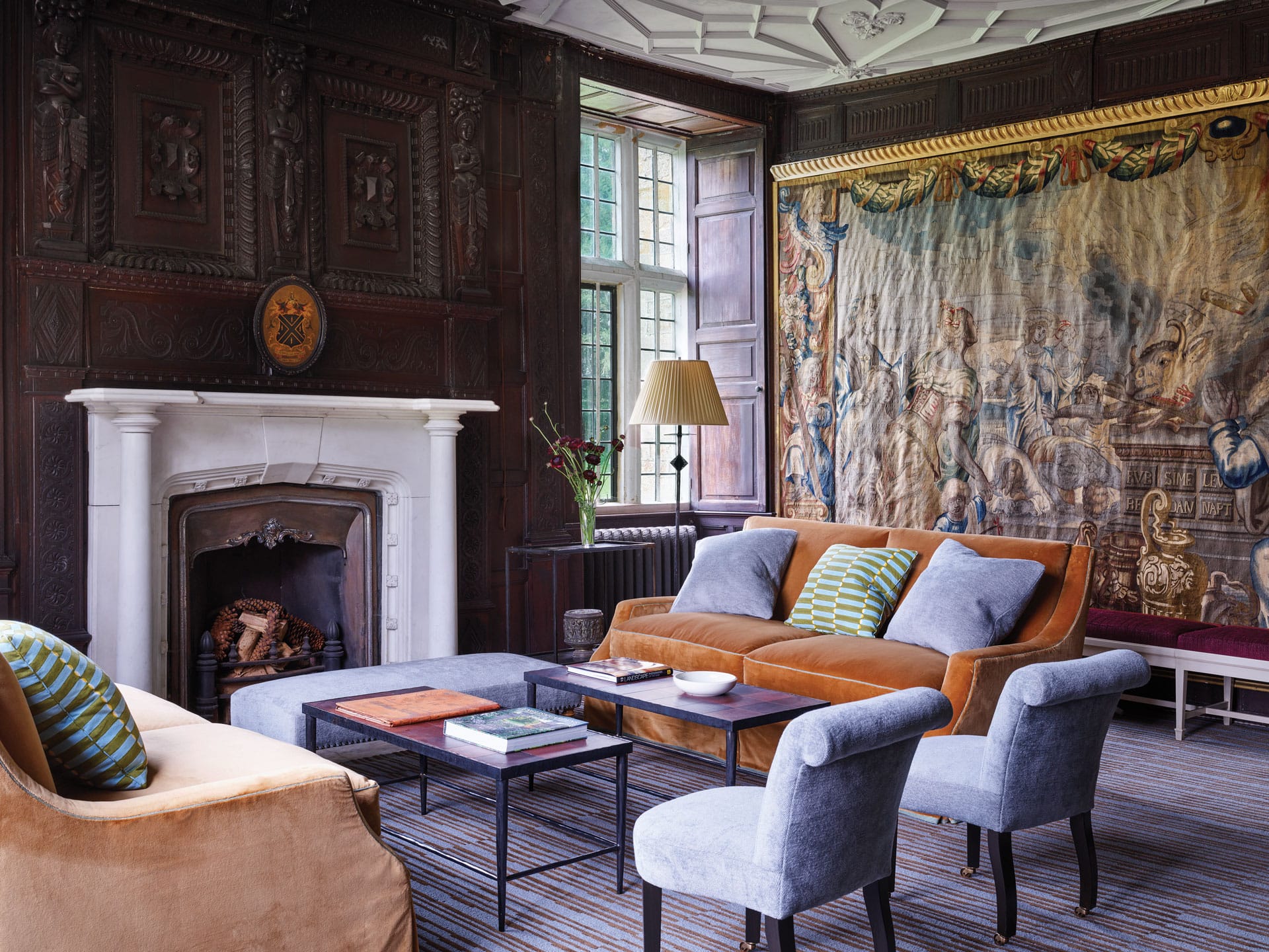 Elegant living room with vintage tapestry, fireplace, orange sofa, blue chairs, striped rug, and large window.