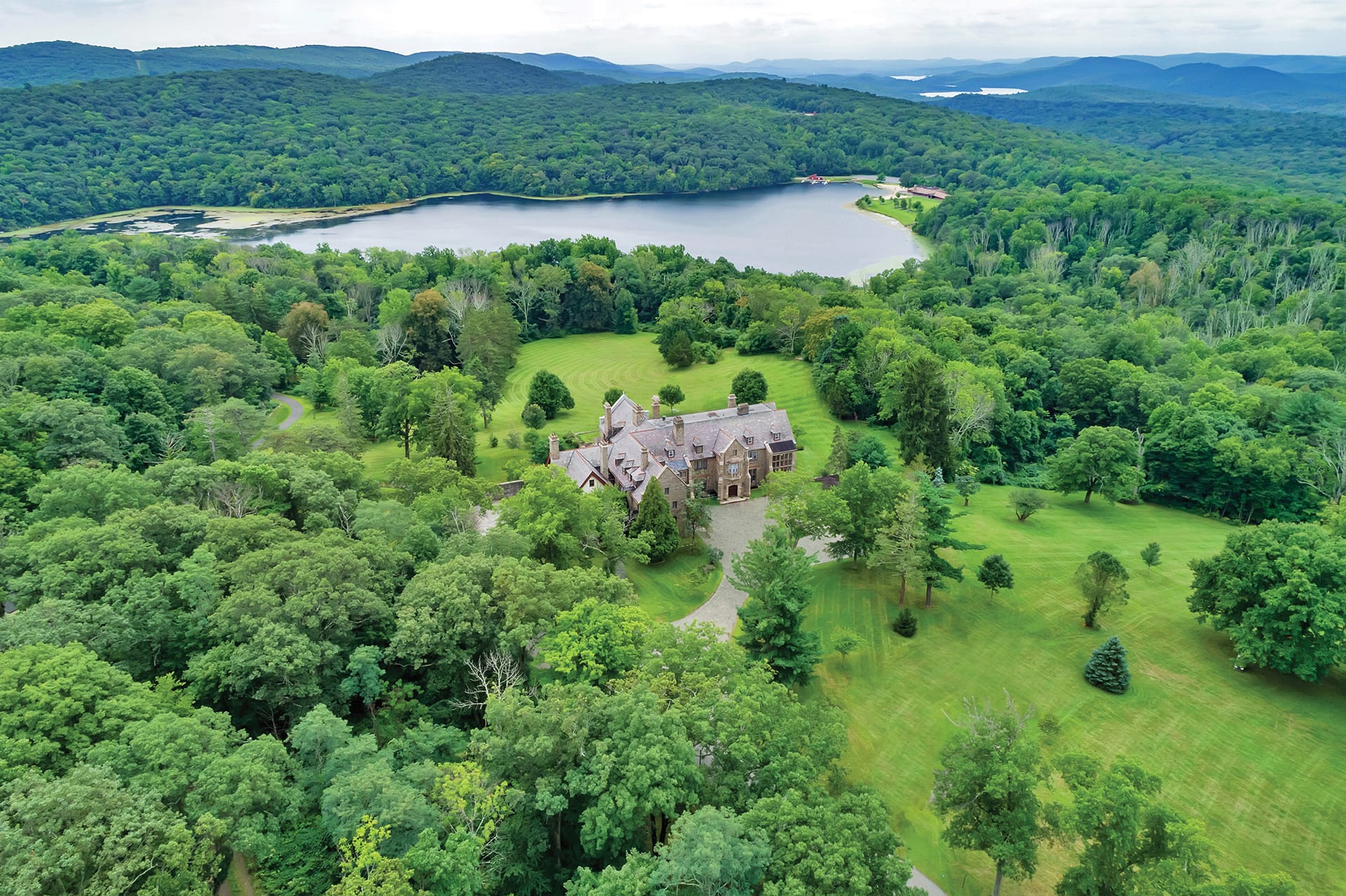 Aerial view of a large mansion surrounded by dense forest with a lake and mountains in the background.