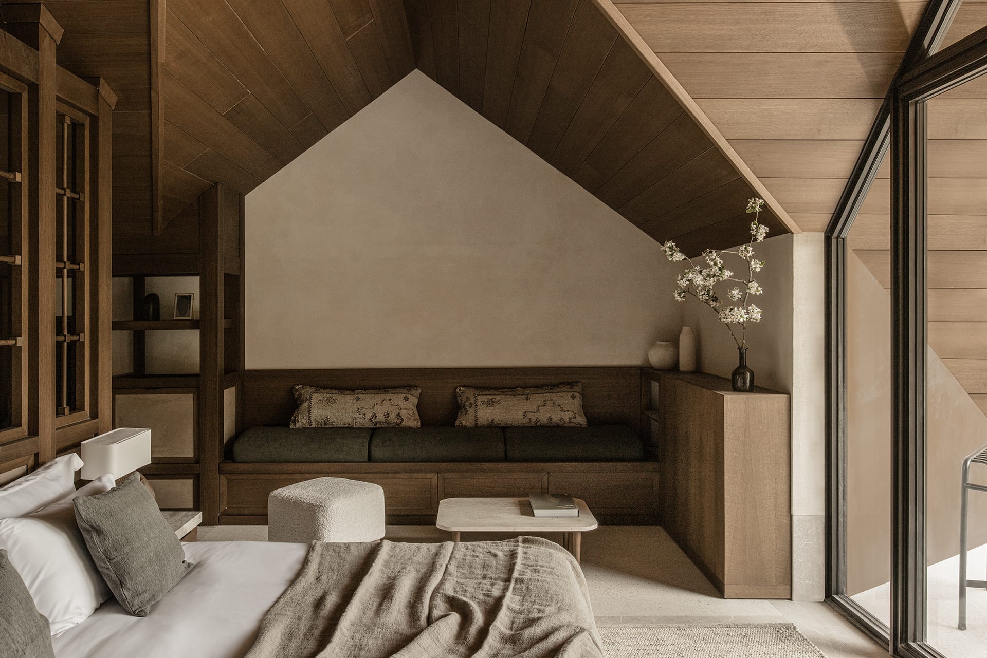 Cozy modern attic bedroom with wooden ceiling, stylish bed, seating area, and large window with soft lighting.