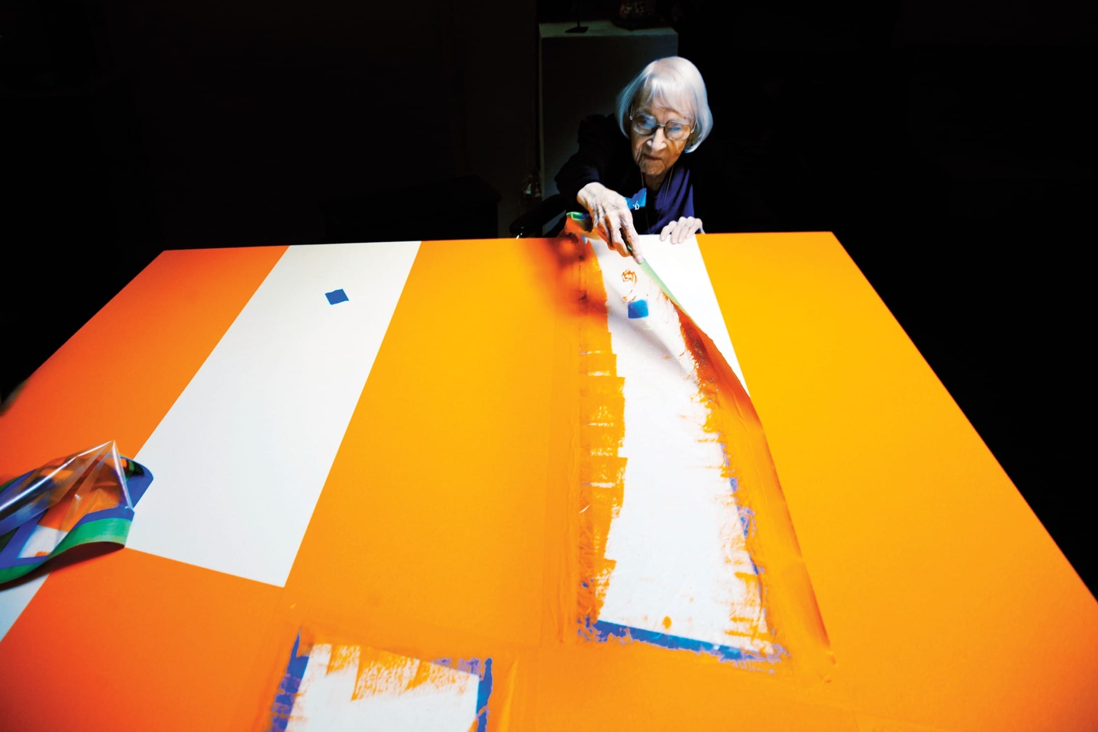 Elderly woman creatively painting a large canvas with bright orange and white colors in an art studio setting.