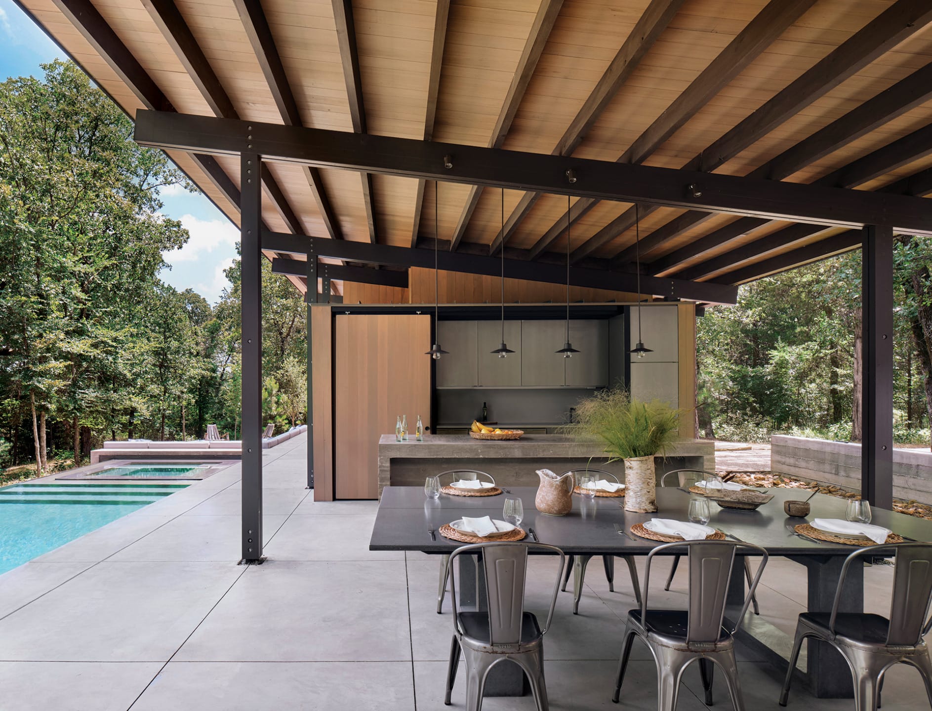 Modern outdoor dining area with a sleek table, chairs, overhead beams, pool, and lush greenery in the background.