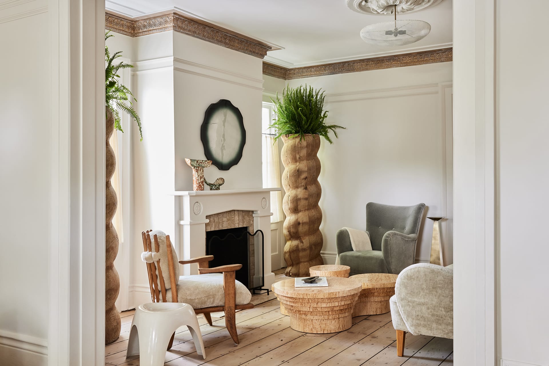 Modern living room with unique wooden chairs, woven side table, fireplace, round mirror, and tall twisted plant holders.