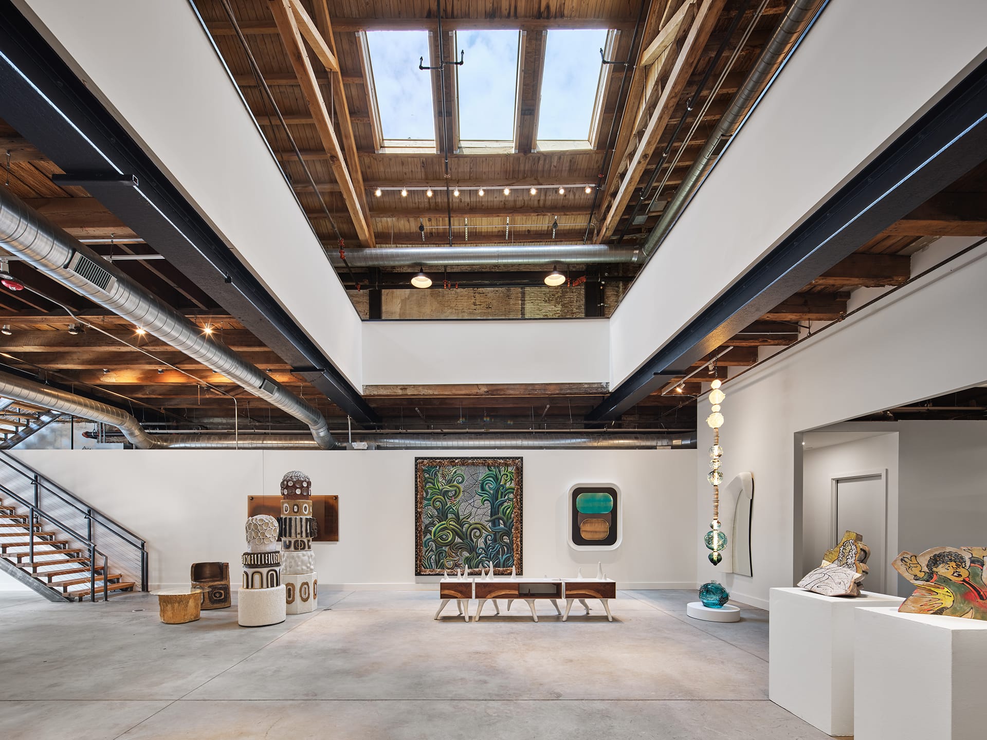 Modern art gallery with various sculptures and paintings, wooden ceiling, skylights, and industrial stairway in open space.
