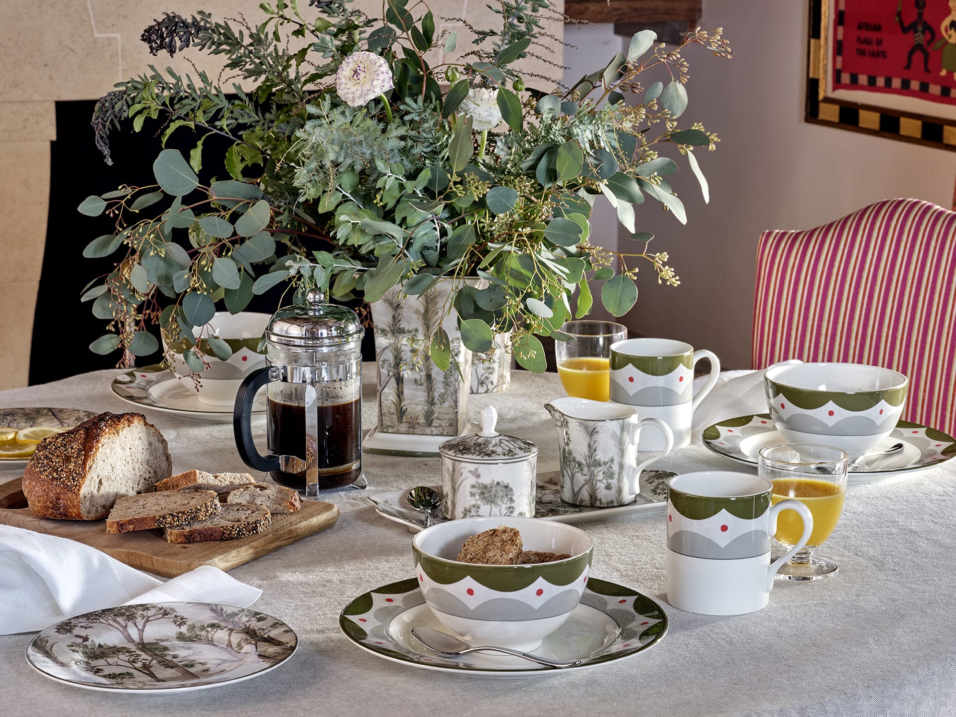 Elegant breakfast table with coffee, assorted breads, orange juice, and floral china, adorned with greenery centerpiece.