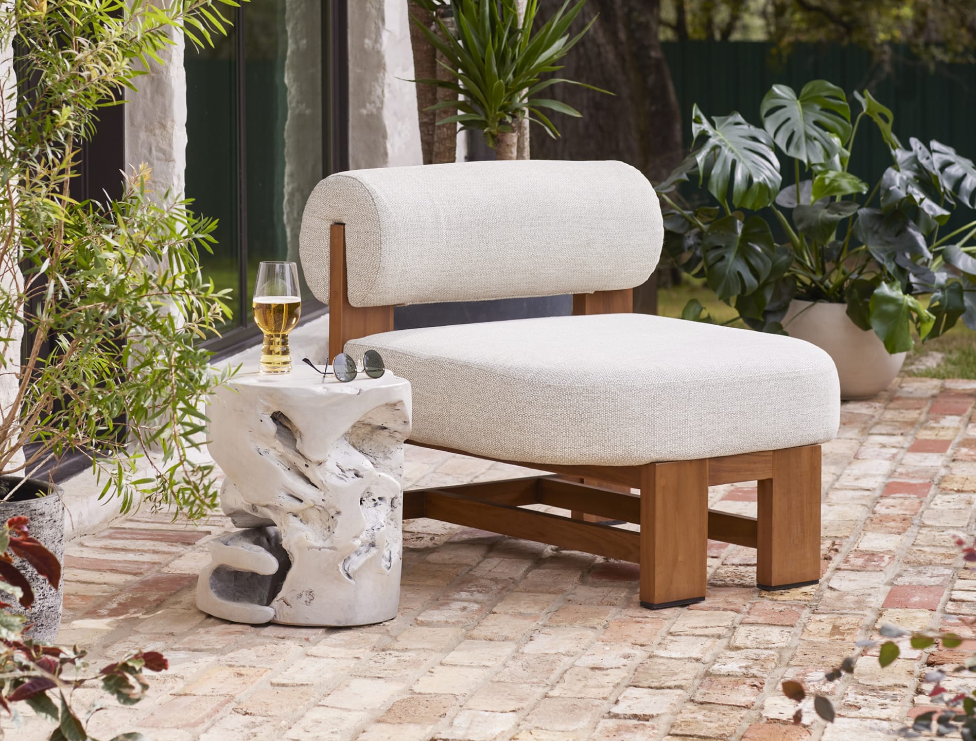 Modern outdoor chair on a brick patio with a rustic side table, drink and sunglasses, surrounded by plants.