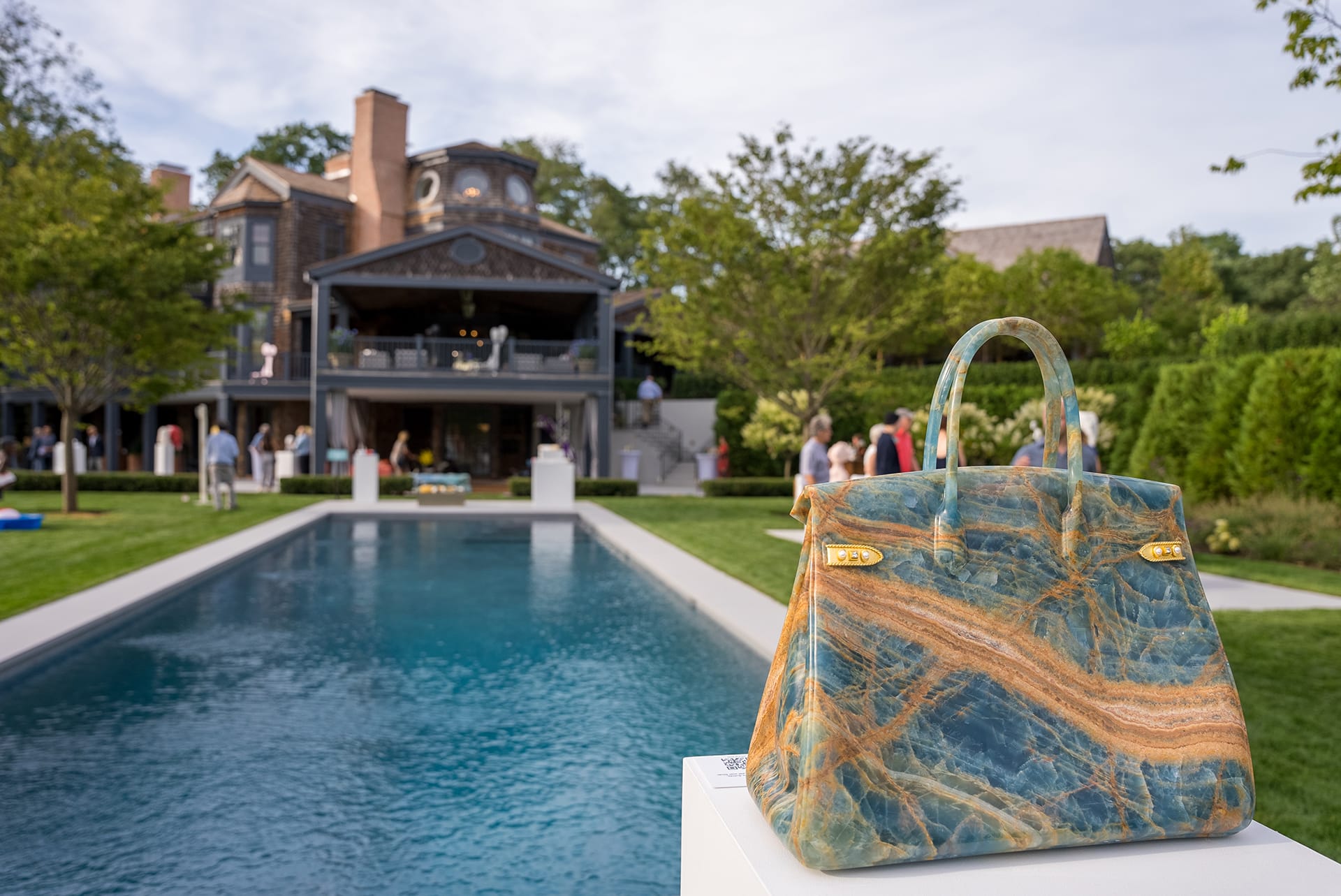 A luxury handbag on display by a poolside with a large house and garden in the background.