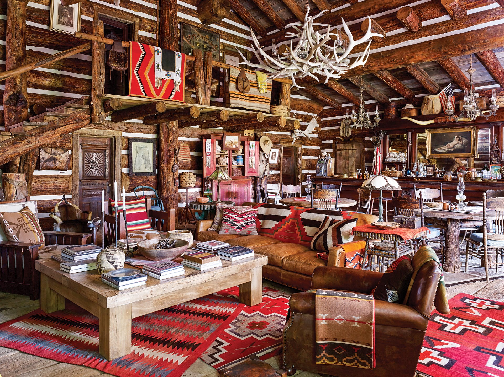 Rustic living room with wooden beams, leather sofa, red patterned rugs, antler chandelier, and a log cabin aesthetic.