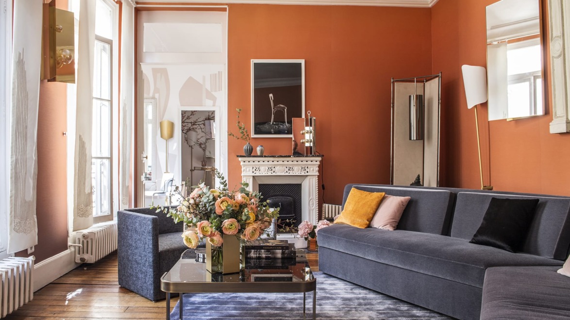 Elegant living room with orange walls, modern furniture, large windows, and a floral centerpiece on a glass coffee table.