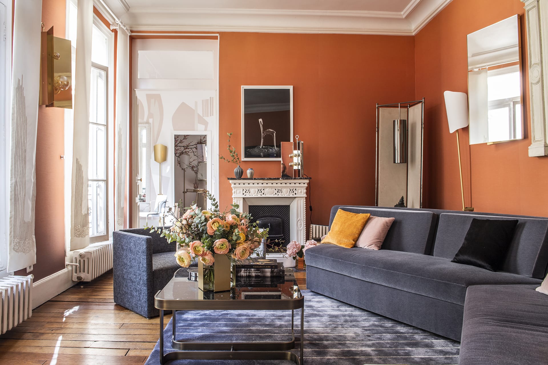 Elegant living room with orange walls, modern furniture, large windows, and a floral centerpiece on a glass coffee table.