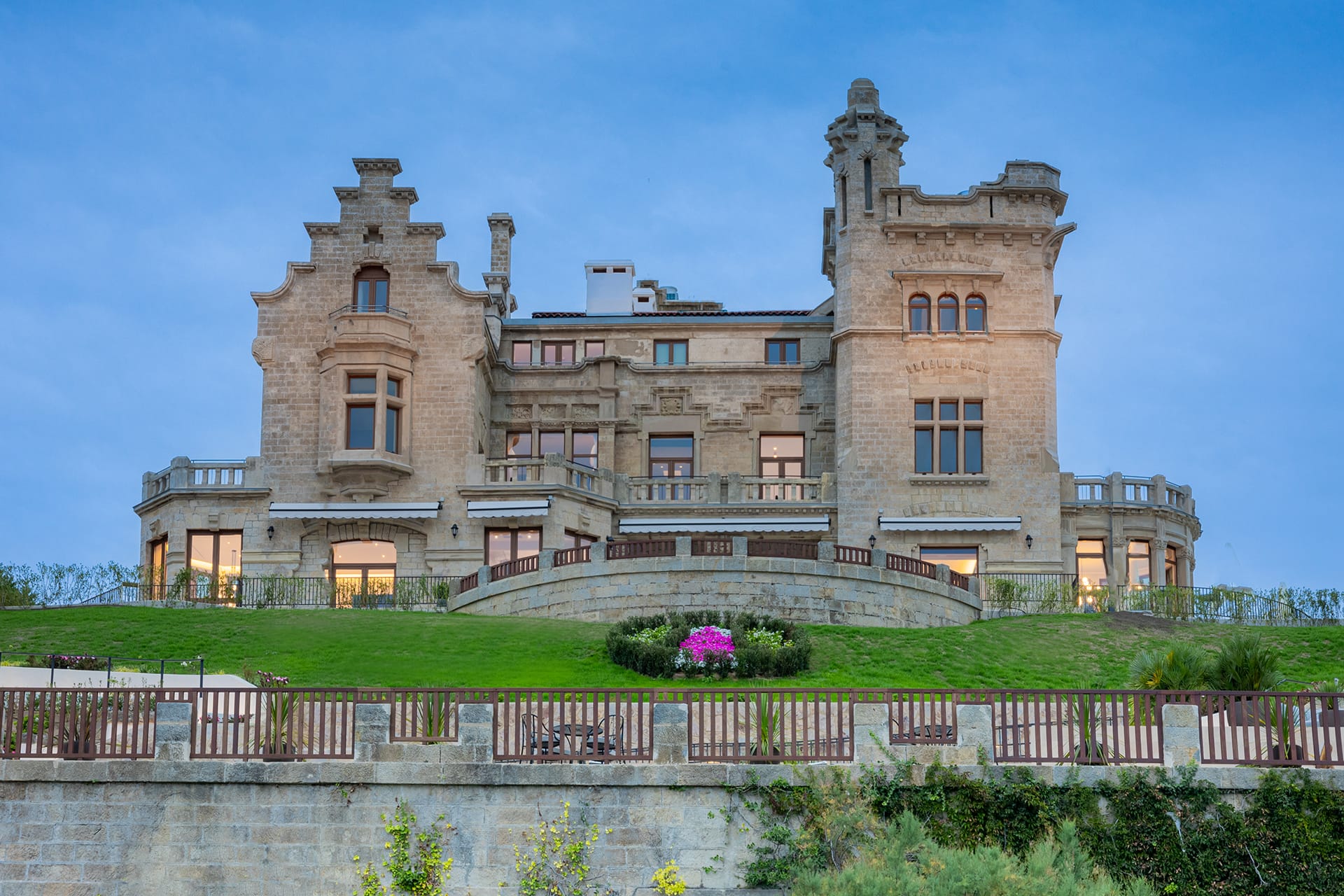 Historic stone castle with intricate architecture, large windows, and a manicured lawn under a clear blue sky.