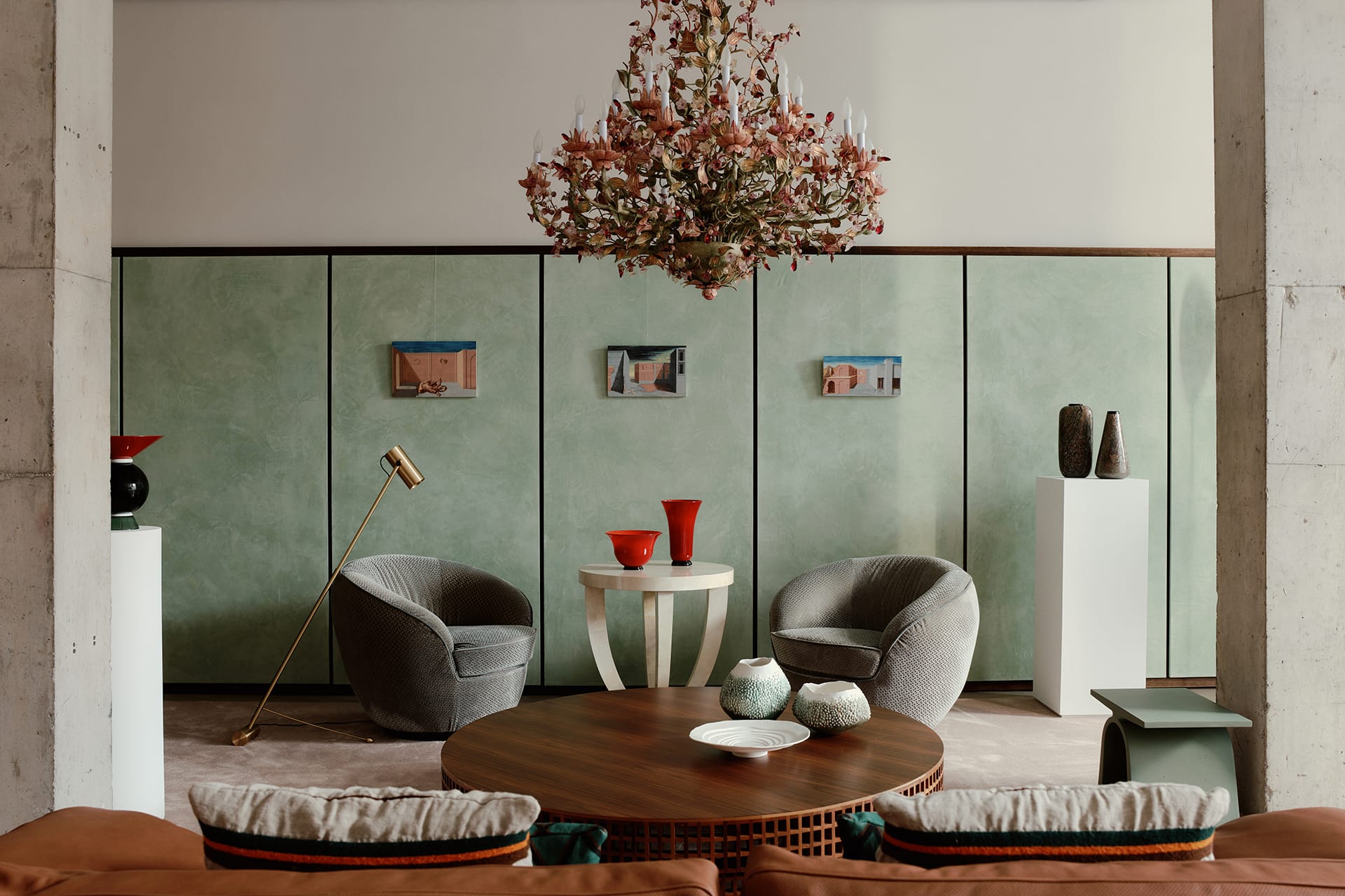 Elegant living room with circular table, two gray armchairs, green walls, floral chandelier, and modern decor elements.
