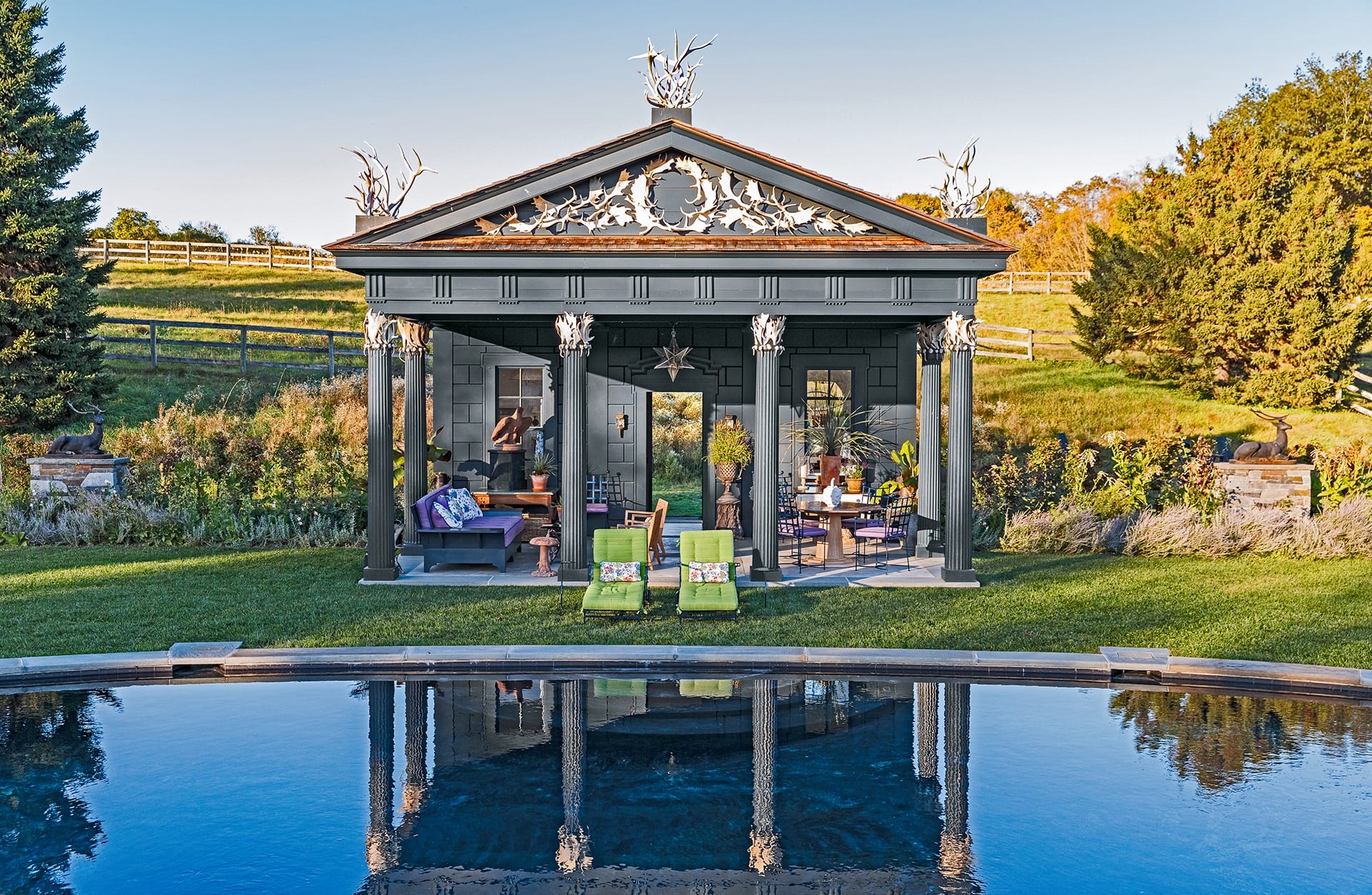 Elegant garden pavilion with rustic decor, seating area, and lawn chairs by a serene pool in a lush green landscape.