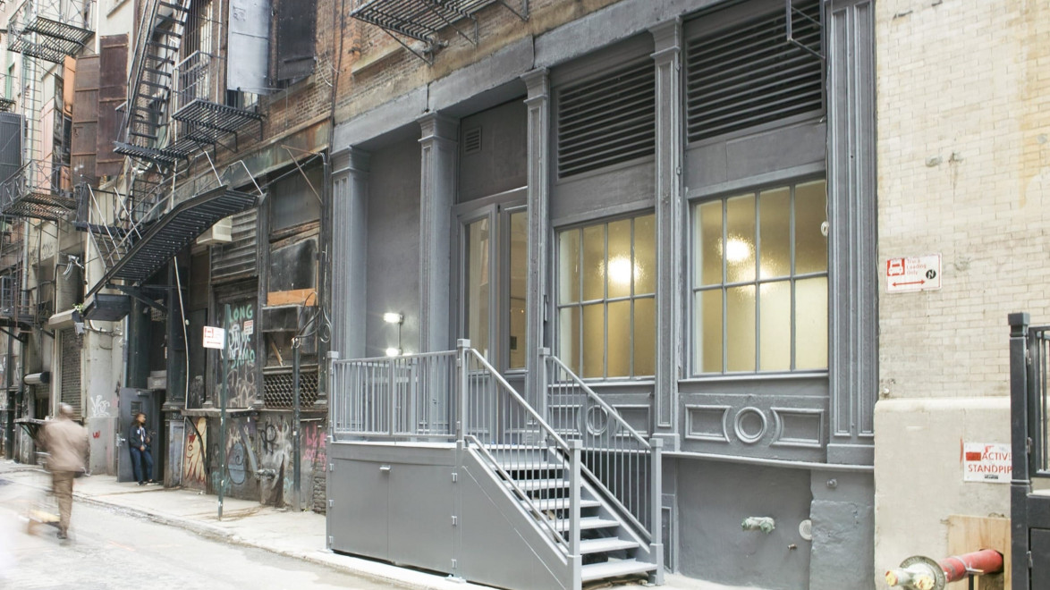 Gray urban building with large windows and a staircase, surrounded by graffiti and fire escapes on a narrow street.