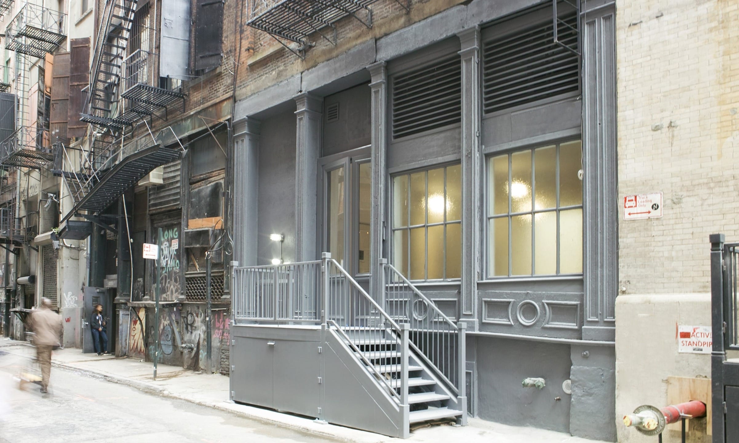 Gray urban building with large windows and a staircase, surrounded by graffiti and fire escapes on a narrow street.