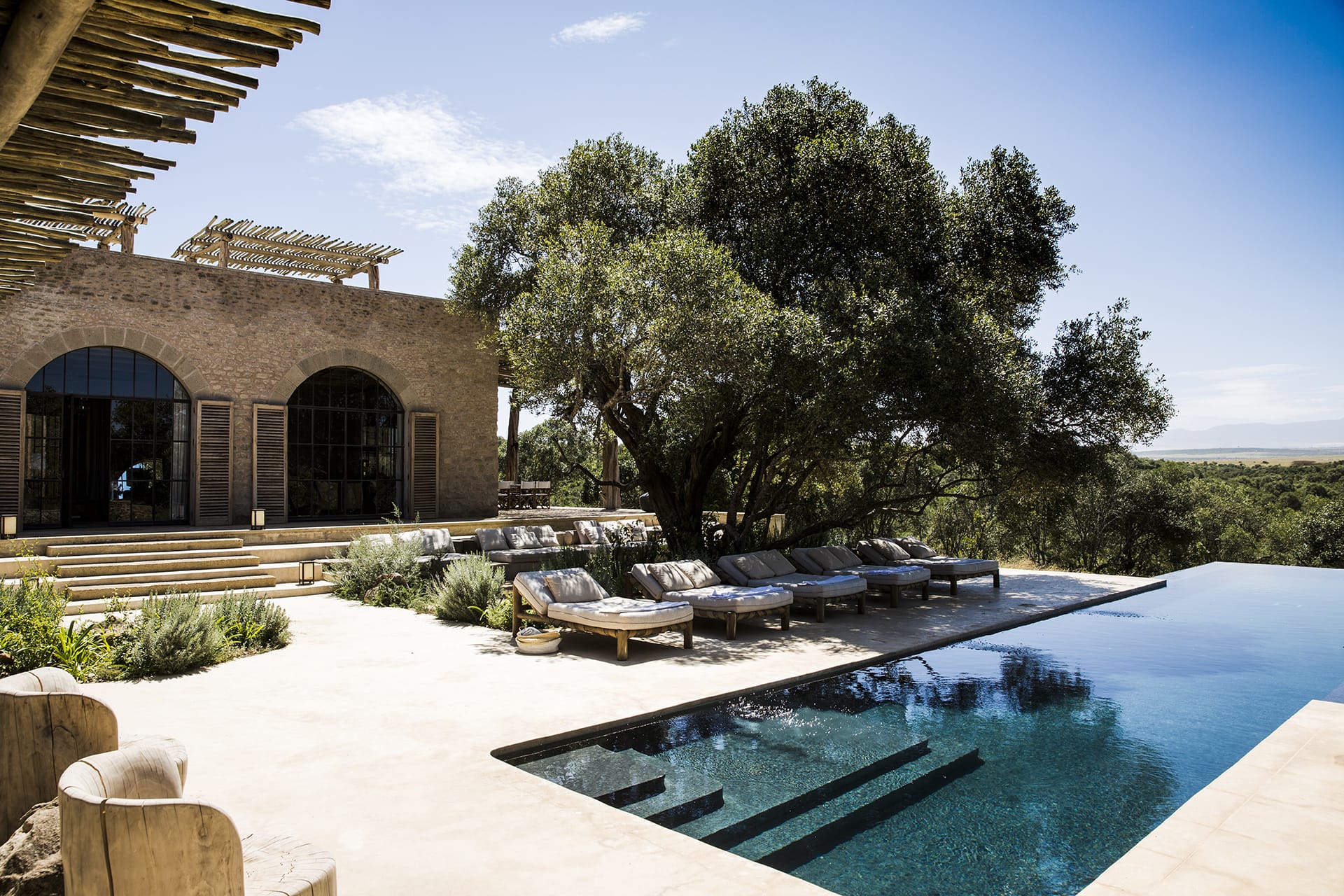 Luxurious villa with sun loungers by an infinity pool, surrounded by greenery under a clear blue sky.