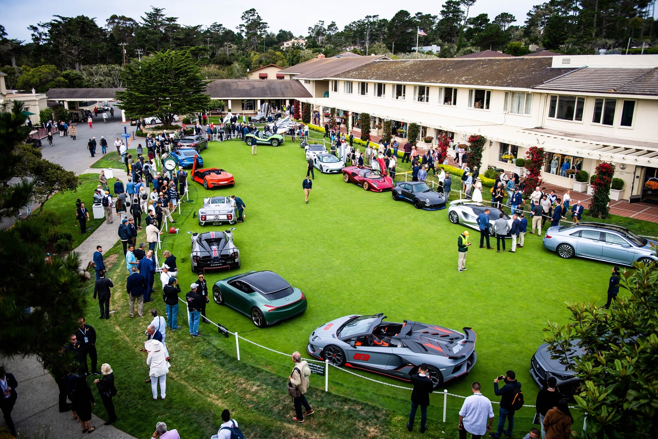 Car show with various luxury vehicles displayed on a green lawn, surrounded by people and a large building.