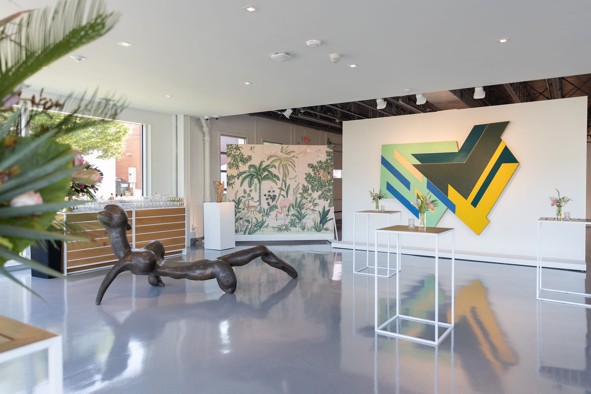 Contemporary art gallery interior with abstract painting, sculpture, and floral decorations on display tables.