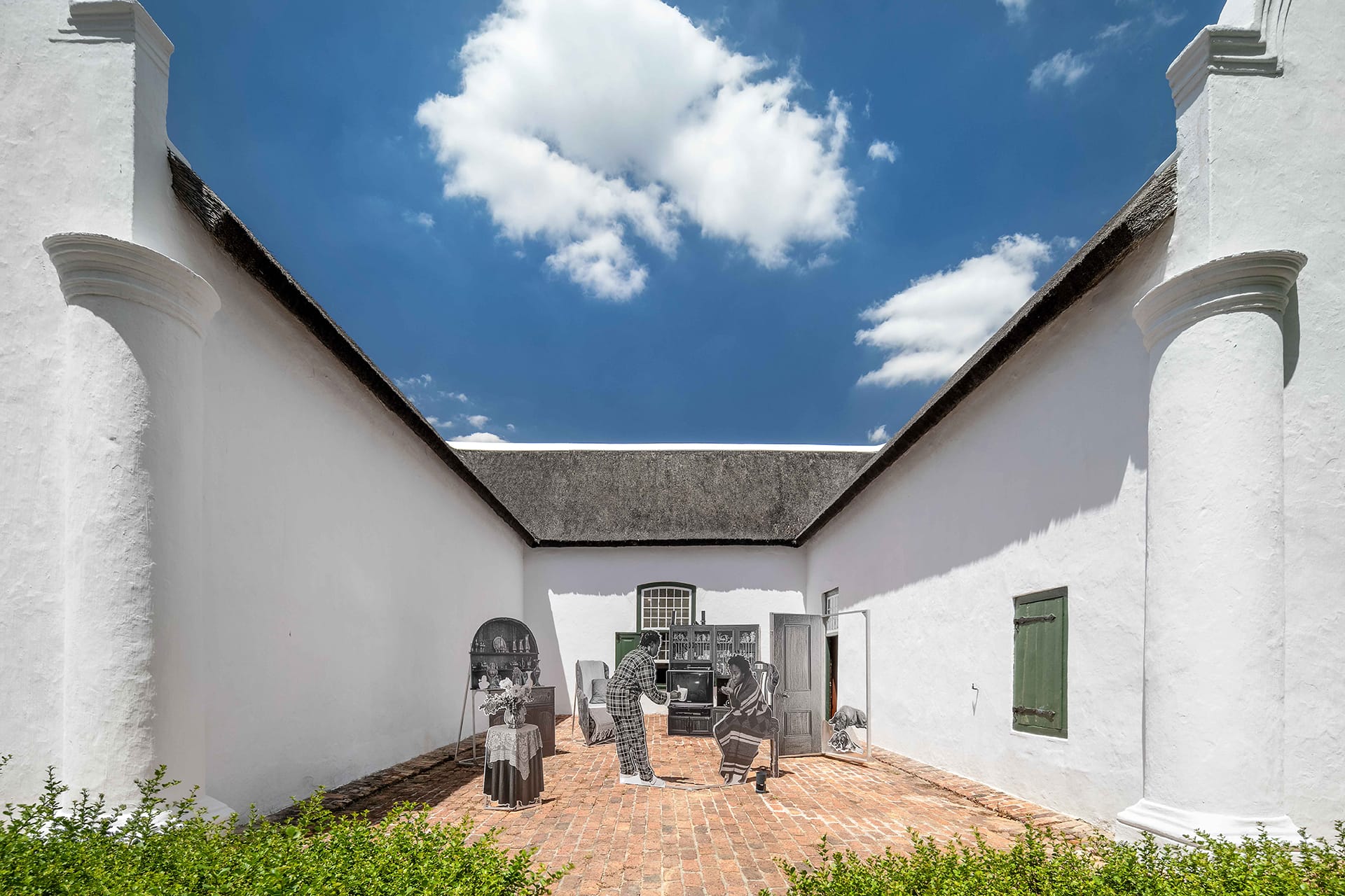 Historic courtyard with thatched roof, blue sky, brick floor, and sculpture of people engaging in daily activities.