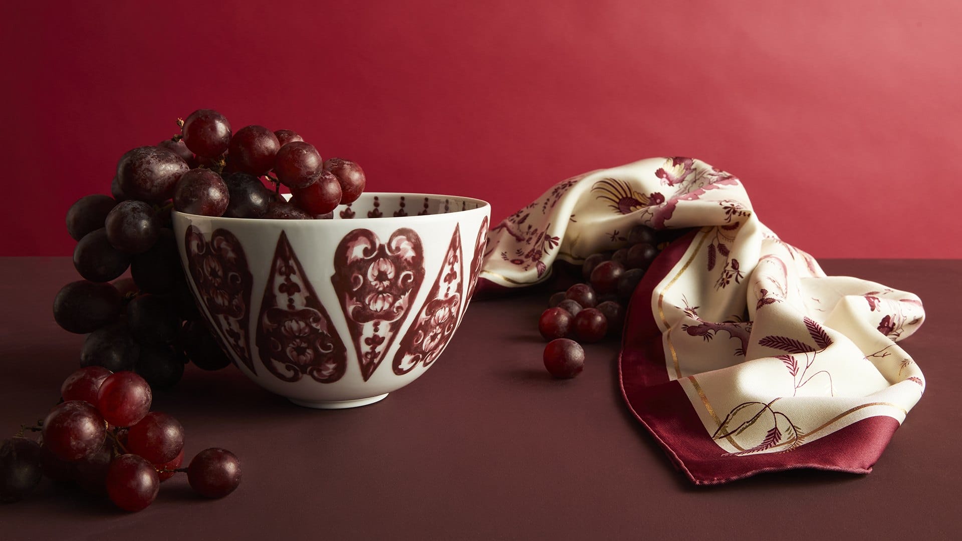 Ornate bowl with red grape design, filled with grapes, beside a patterned cloth on a burgundy background.