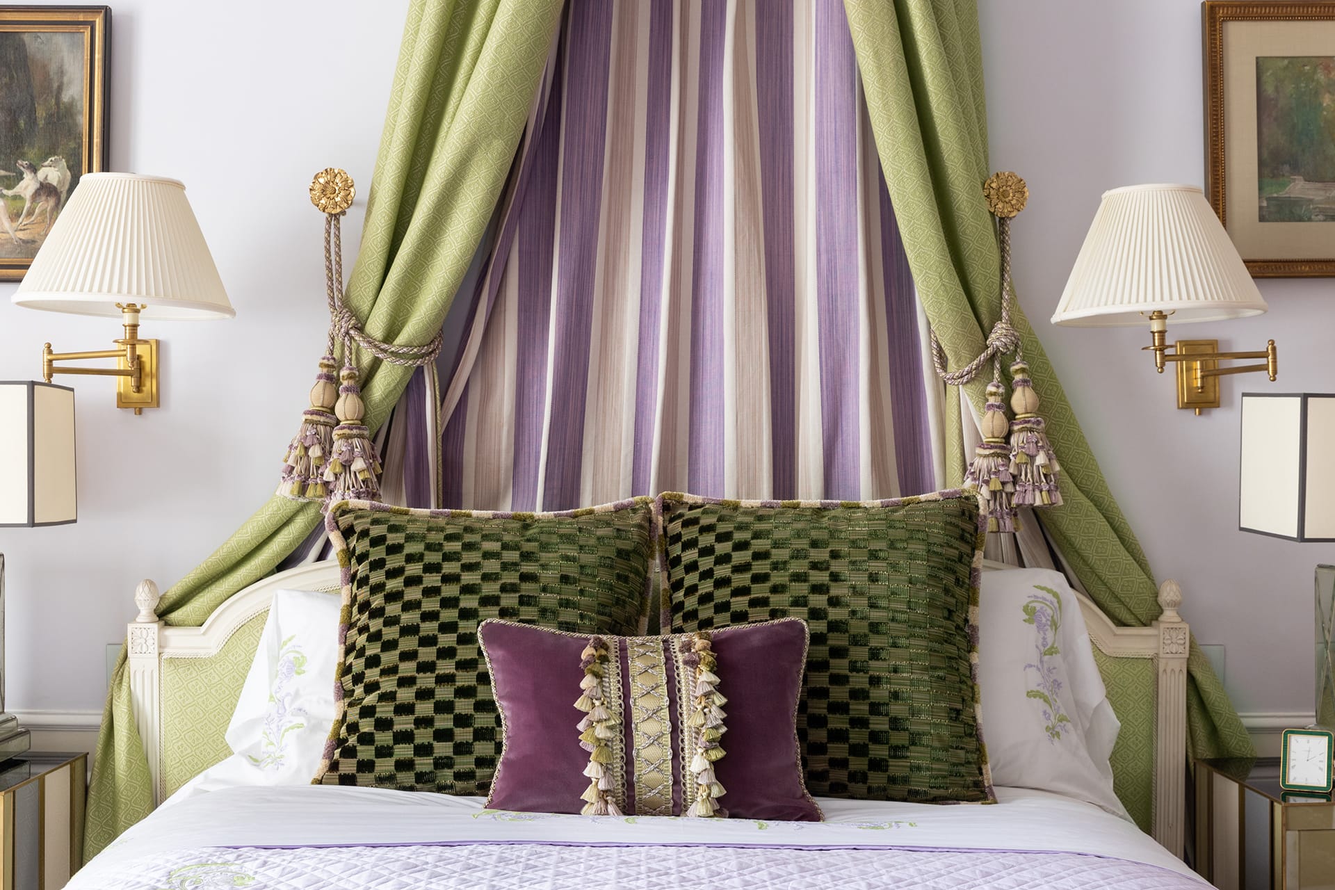 Elegant bedroom with canopy bed, green and purple pillows, decorative curtains, wall lamps, and framed artwork.