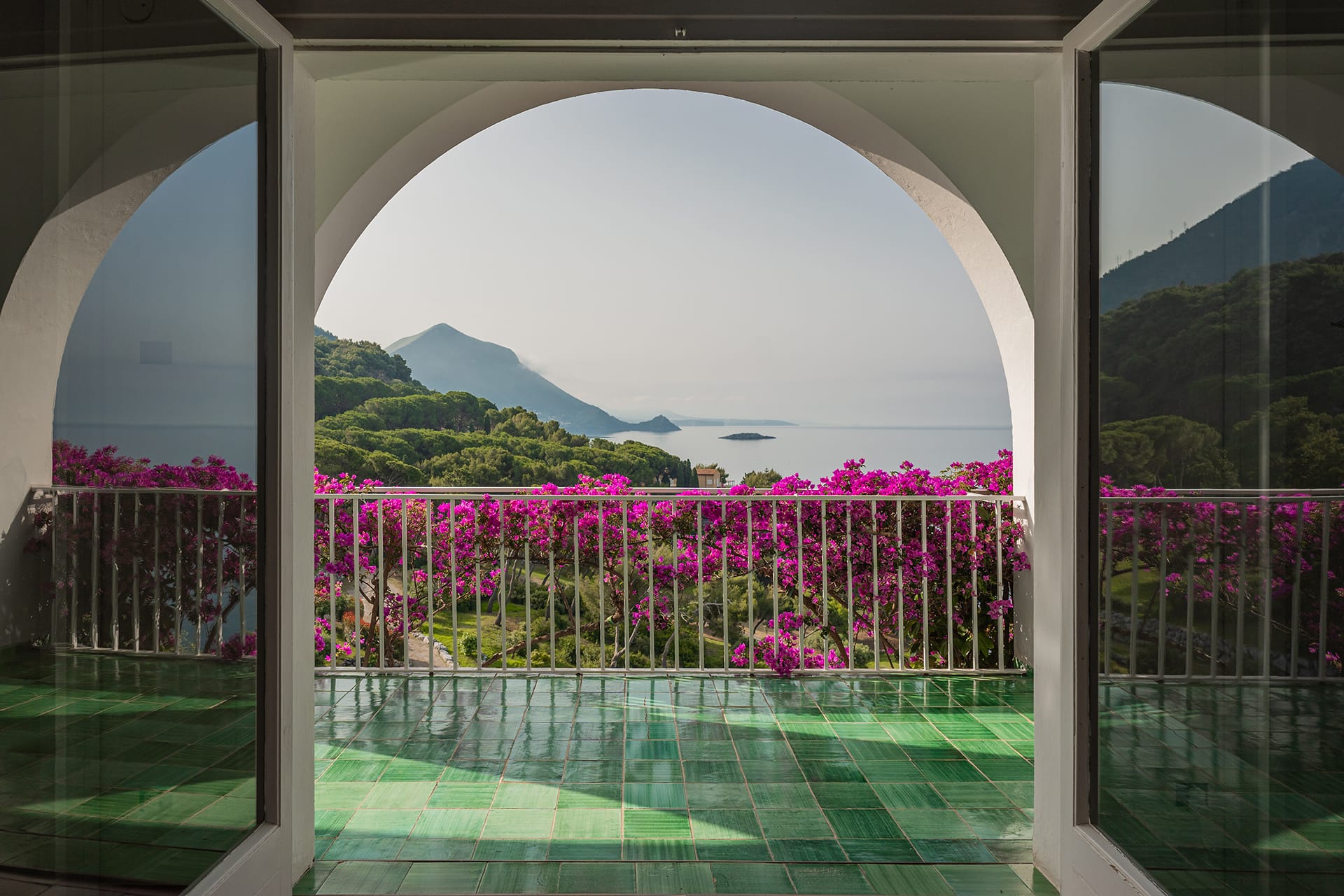 View from an arched balcony overlooking a serene sea and lush mountains, framed by vibrant purple bougainvillea flowers.