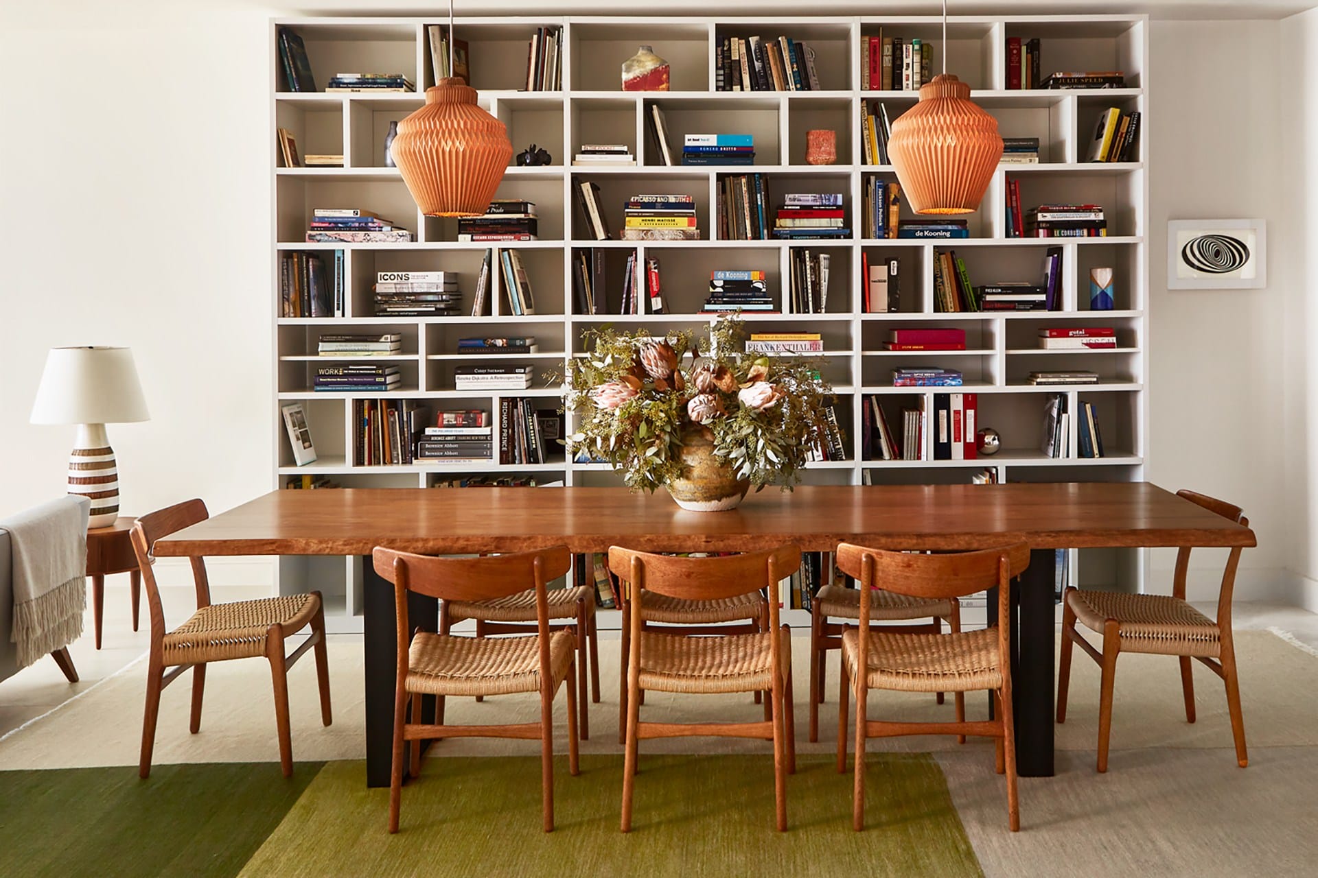 Wooden dining table with six chairs, green rug, and a bookshelf filled with books and decor items in the background.