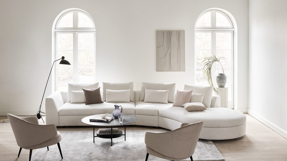 Modern living room with a curved white sofa, round coffee table, two armchairs, large windows, and minimalist decor.