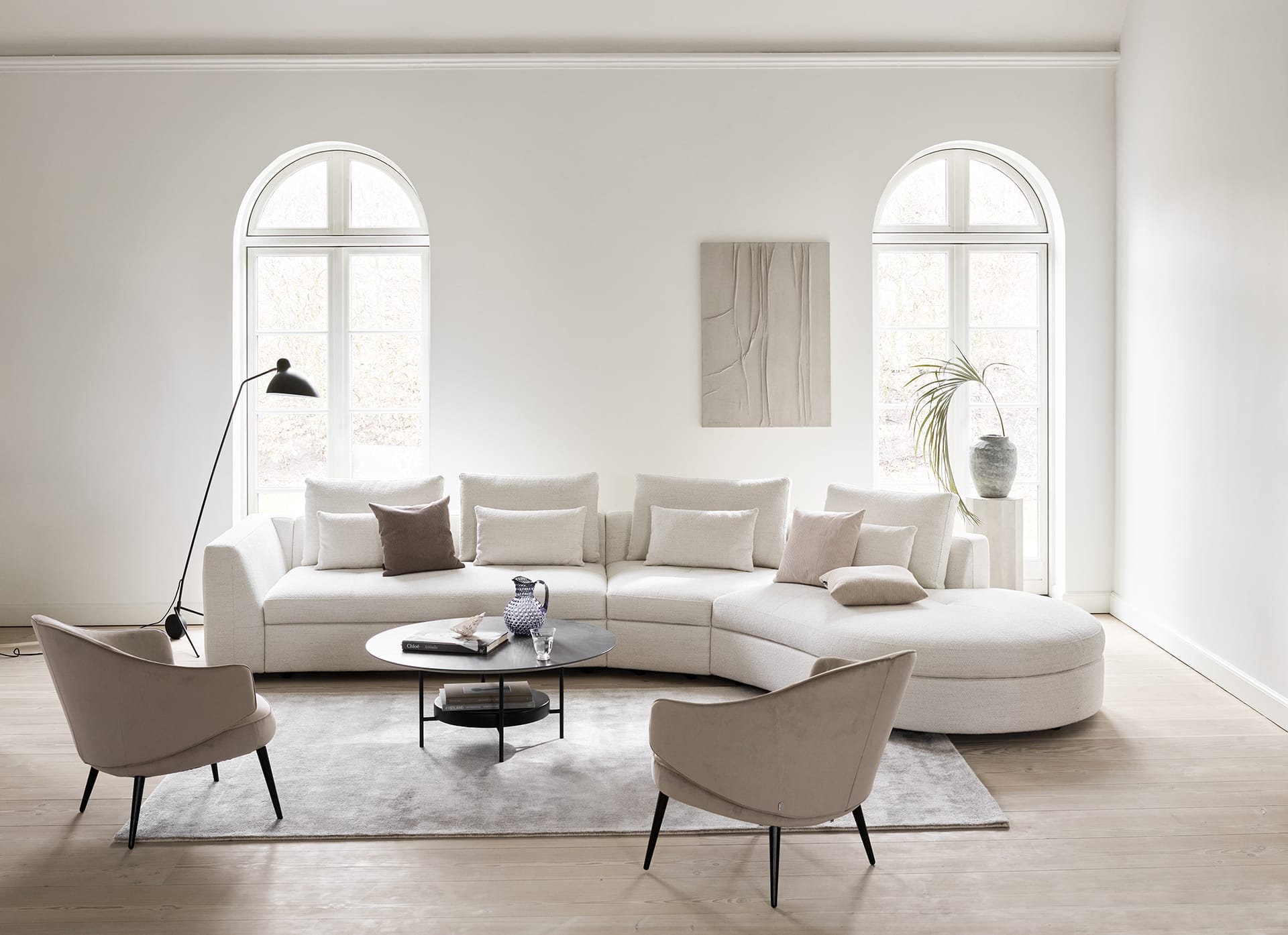 Modern living room with a curved white sofa, round coffee table, two armchairs, large windows, and minimalist decor.