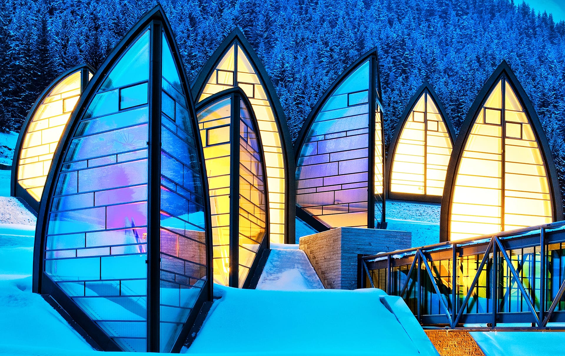 Modern, illuminated glass chapel structures in a snowy mountain setting at dusk with a bridge in the foreground.