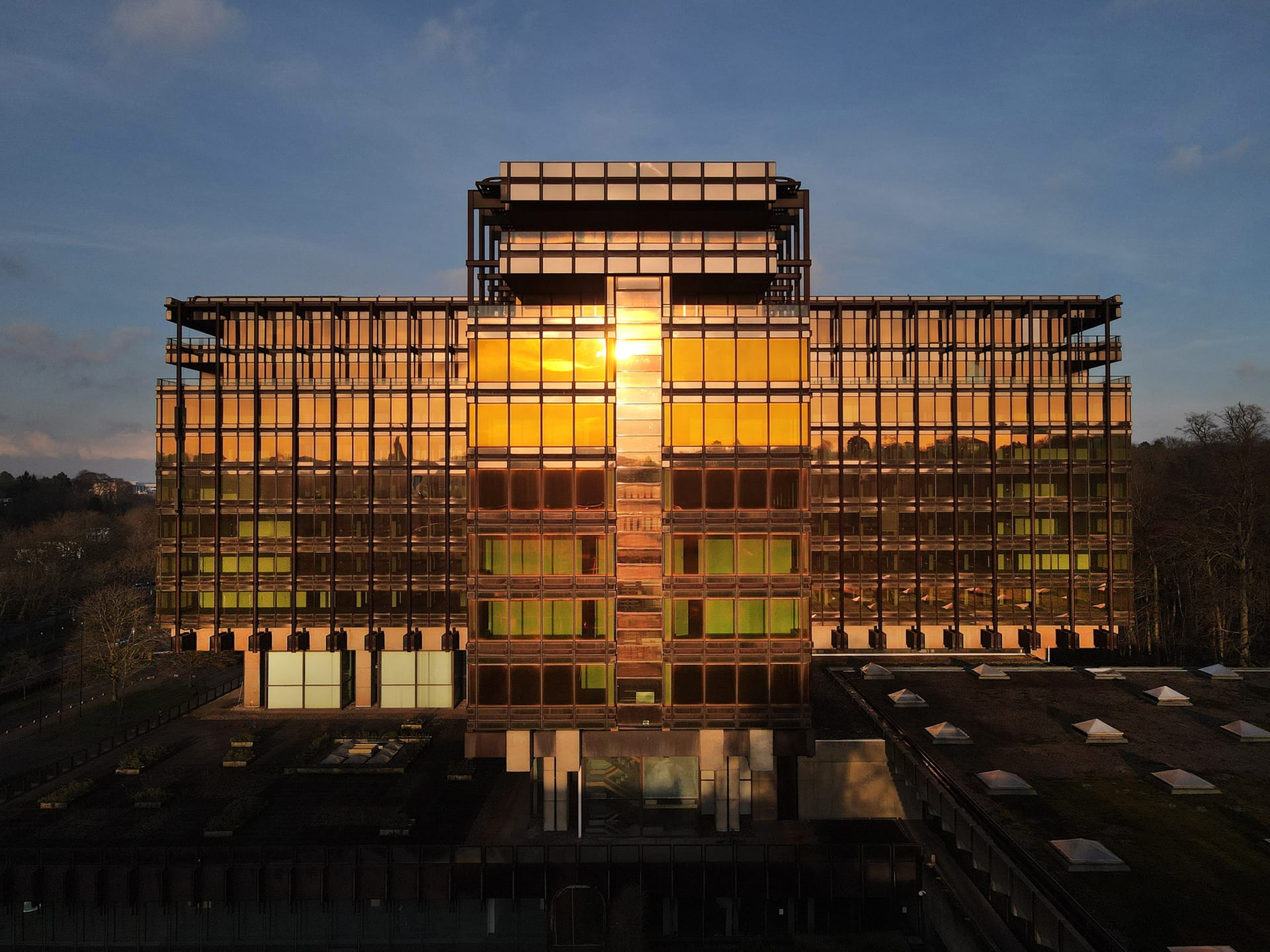 Modern glass building reflecting sunlight with a backdrop of trees and a clear sky.