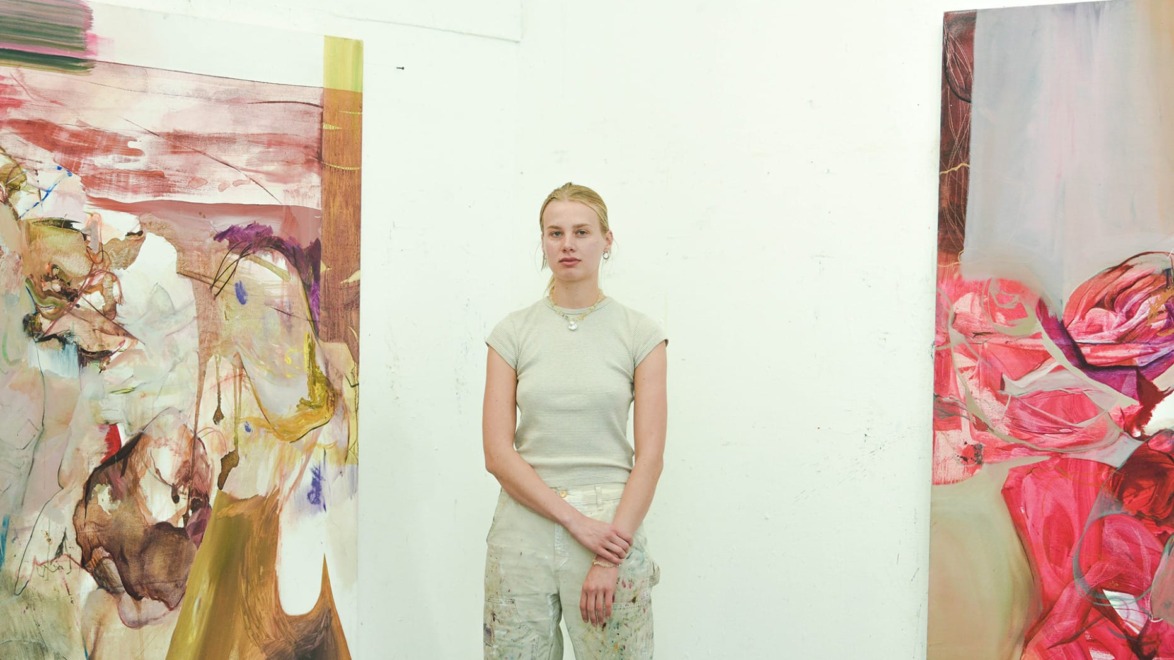 Artist standing in studio between two abstract paintings on a white wall.