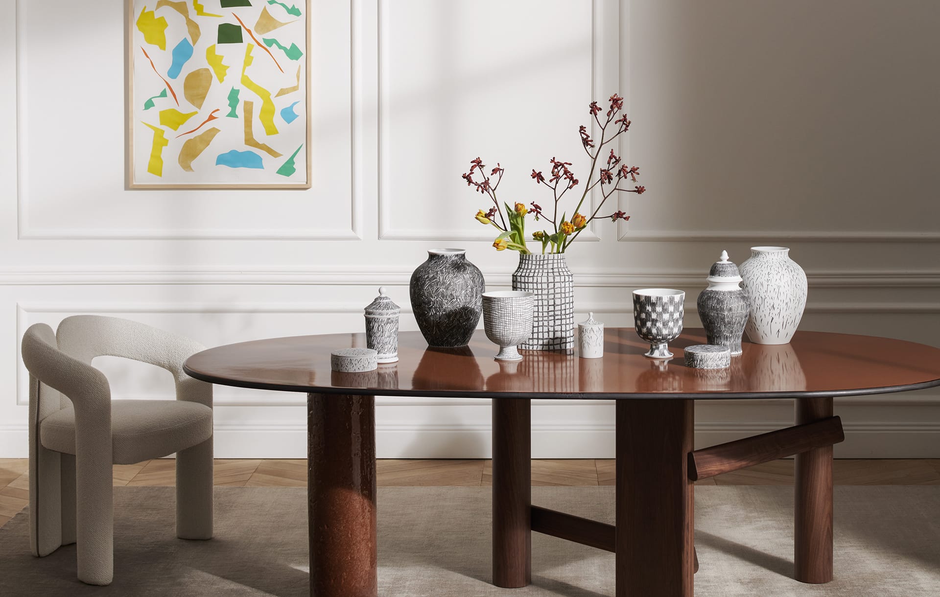 Modern dining room with a wooden table displaying various decorative vases, an abstract wall painting, and a chair.