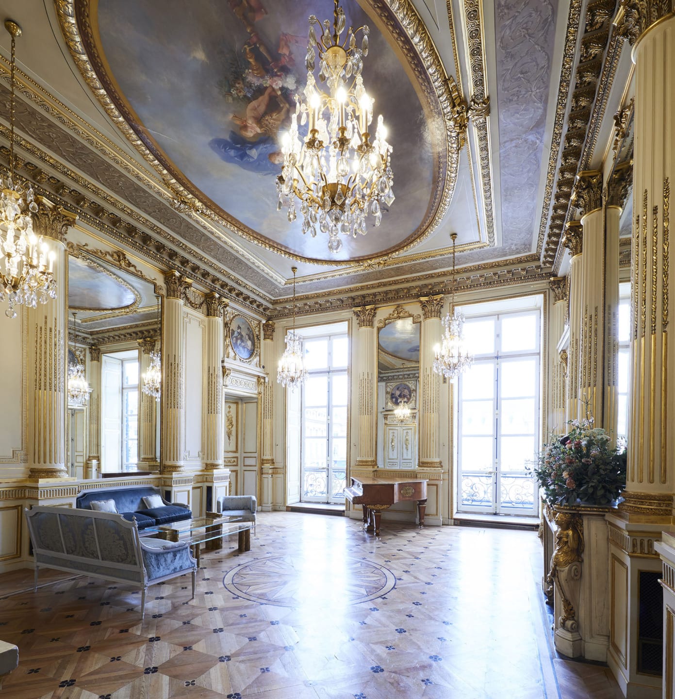 Ornate historical room with chandeliers, frescoed ceiling, large mirrors, and luxurious furniture with natural light streaming in