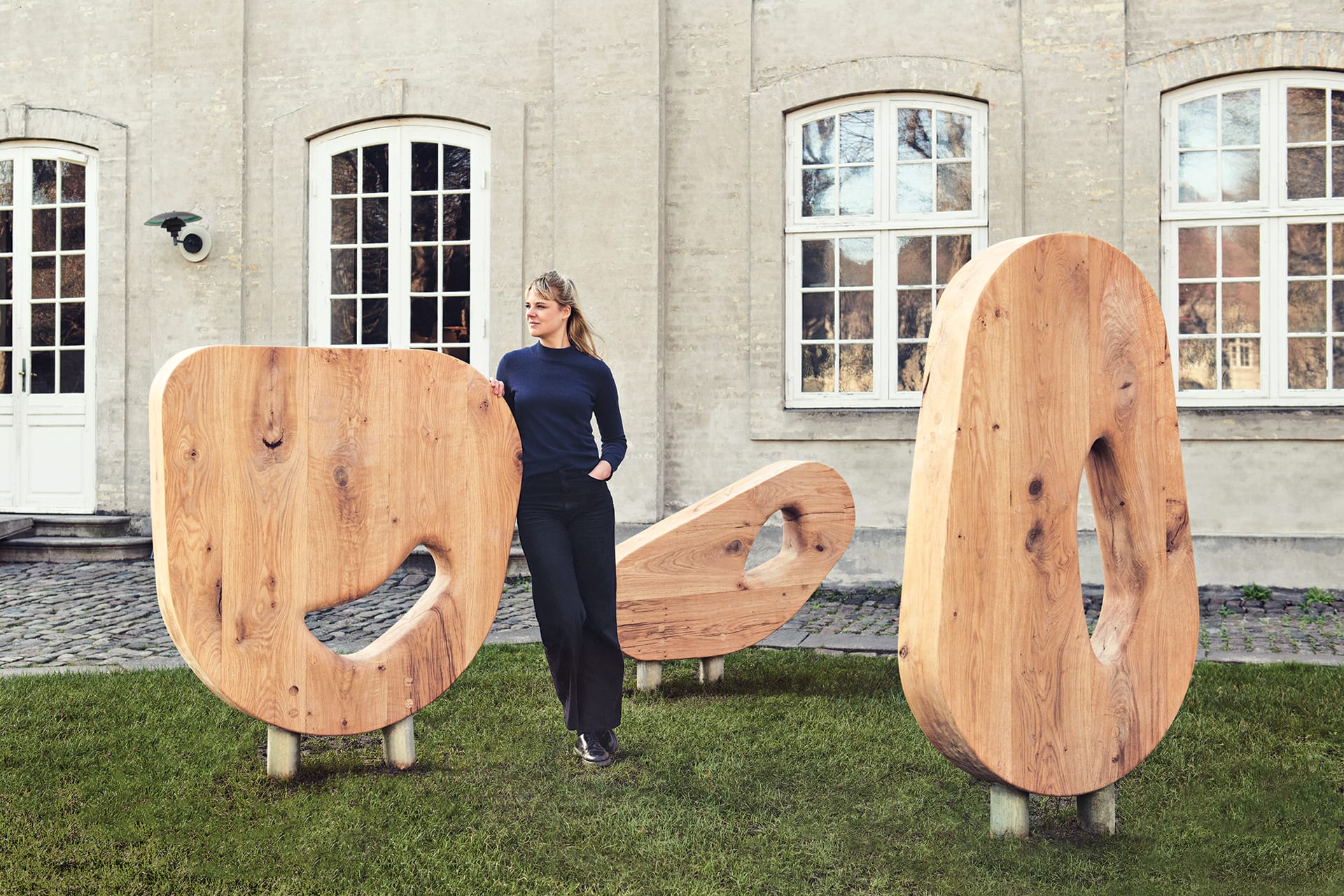 Woman standing beside large abstract wooden sculptures in outdoor courtyard setting.