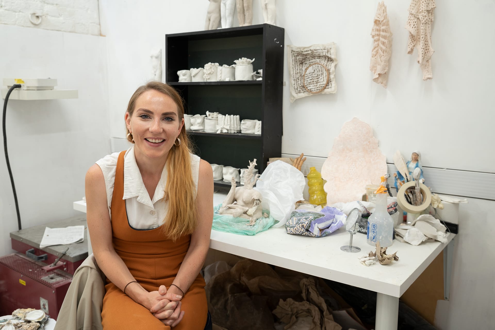 Artist sitting and smiling in a studio with sculptures and art materials on a desk behind her.