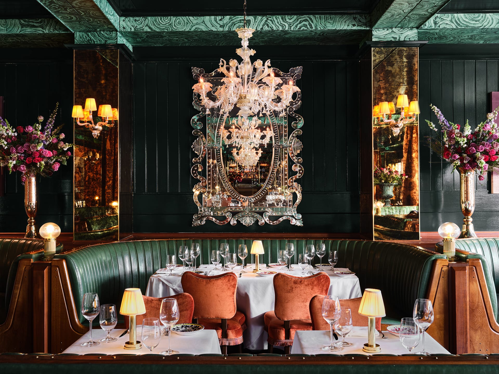 Elegant restaurant interior with chandelier, green booths, floral arrangements, mirrors, and table settings.