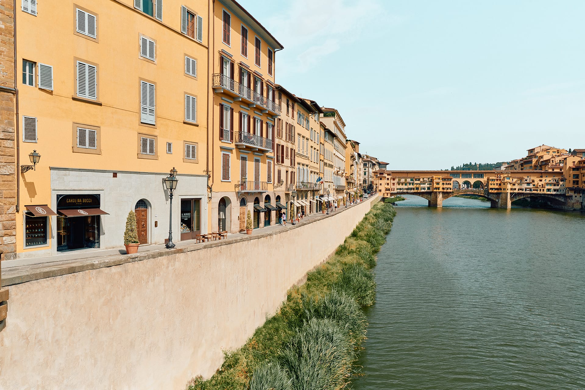 Riverside view of historic buildings and Ponte Vecchio bridge over the Arno River in Florence, Italy, under a clear sky.
