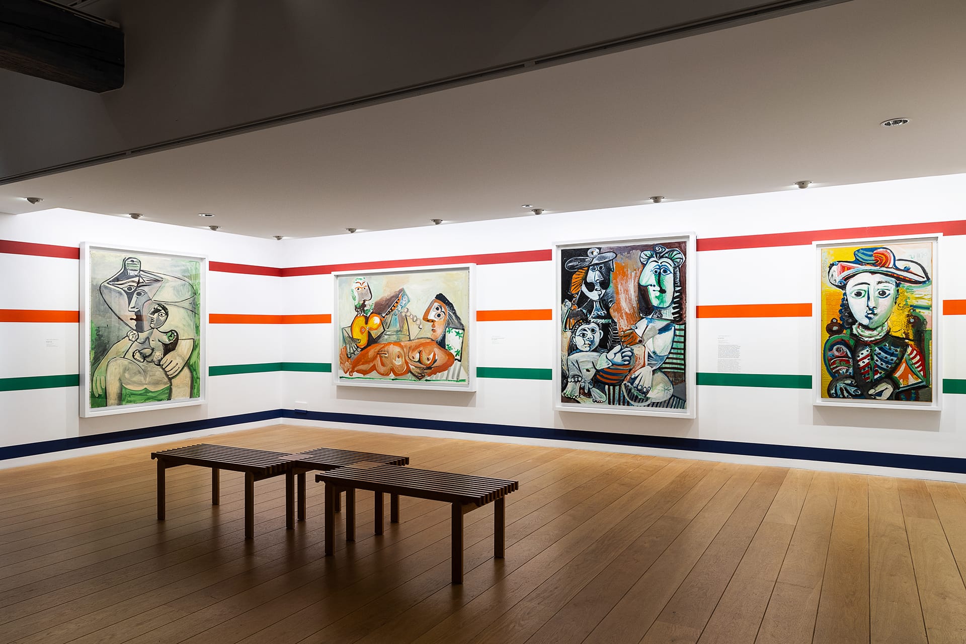 Art gallery interior with four colorful abstract paintings on the wall and two wooden benches on the wooden floor.