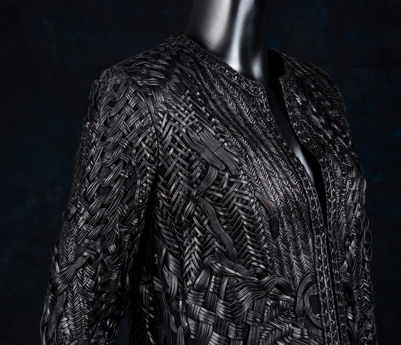 Intricately woven black garment on a mannequin against a dark background, showing detailed craftsmanship and texture.